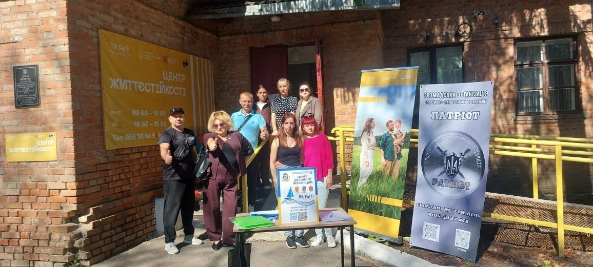 Група людей урочисто позує для спільного фото на вулиці біля входу до цегляної будівлі Координаційного центру «Вільні разом» у Кропивницькому, де в межах всебічної підтримки переселенців з Херсонщини було укладено меморандуми про співпрацю з ГО «Спортивно-патріотична організація «Патріот» та БО «100% життя. Кропивницький». Учасники робочої зустрічі вишикувалися на ґанку та біля жовтого пандуса: на передньому плані ліворуч стоять чоловік у чорній кепці й тактичному одязі та жінка у бордовому костюмі й сонцезахисних окулярах, які показують жест схвалення, а по центру біля столу з інформаційною табличкою стоять дві дівчини. Ліворуч на цегляній стіні закріплено великий жовтий банер «Центр життєстійкості» з графіком роботи та номером телефону [+380 66 356 4991], а праворуч встановлено мобільний стенд організації «ПАТРІОТ» із символікою тризуба, що відображає консолідацію зусиль задля допомоги людям.