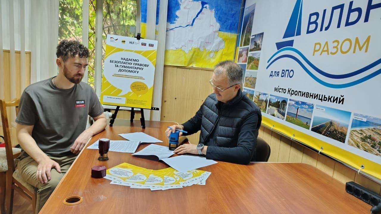 Двоє чоловіків сидять за великим дерев'яним столом у кабінеті Координаційного центру «Вільні разом» у Кропивницькому під час завірення документів автоматичним штампом у межах спільної роботи з БО «100% життя. Кропивницький» задля всебічної підтримки переселенців з Херсонщини. Ліворуч за столом сидить представник благодійної організації у сірій футболці, який спостерігає за процесом, а праворуч — чоловік в окулярах та чорній жилетці завіряє підписані папери угод поруч із розкладеними жовтими буклетами. Кадр зроблено на тлі банера «ВІЛЬНІ РАЗОМ для ВПО місто Кропивницький» із фотоілюстраціями херсонських краєвидів та плаката про надання правової допомоги, що відображає організацію прийомів мобільної команди для оцінки індивідуальних потреб, проведення консультацій та видачі сертифікатів 70 особам.