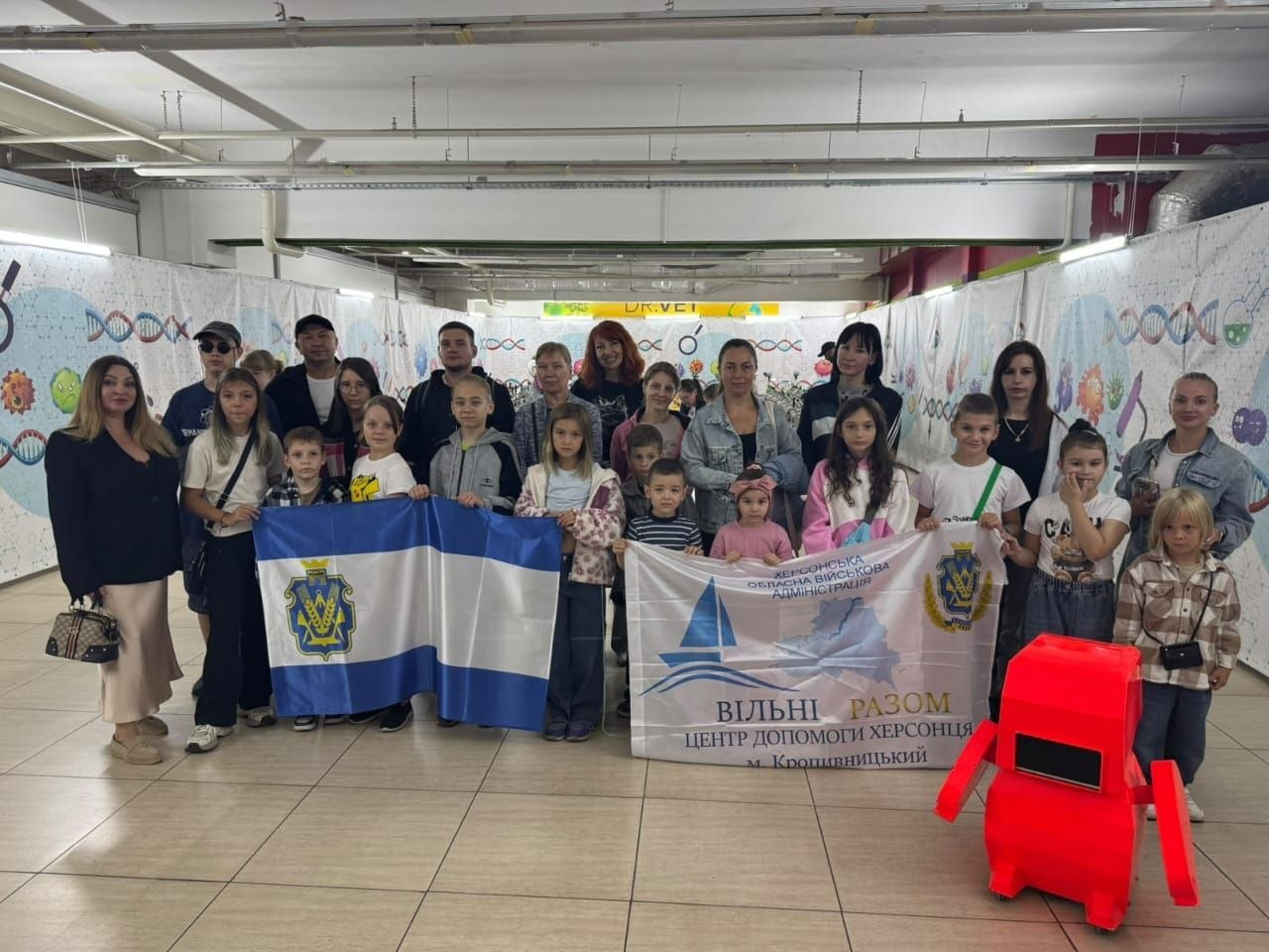Велика група дітей та дорослих із числа переселенців з Херсонщини урочисто позує для спільного фото в просторому залі, де на передньому плані праворуч стоїть велика червона іграшкова фігура робота. У першому ряду ліворуч діти тримають прапор Херсонської області з офіційним гербом у центрі, а праворуч інші учасники тримають великий білий банер хабу з написом «Херсонська обласна військова адміністрація. ВІЛЬНІ РАЗОМ. Центр допомоги херсонцям м. Кропивницький». Усі присутні вишикувалися на світлій кахельній підлозі на тлі довгих білих декорацій, прикрашених яскравими малюнками, що відображає роботу в межах всебічної підтримки від Координаційного центру та ілюструє згуртованість та підтримку херсонських родин в евакуації.