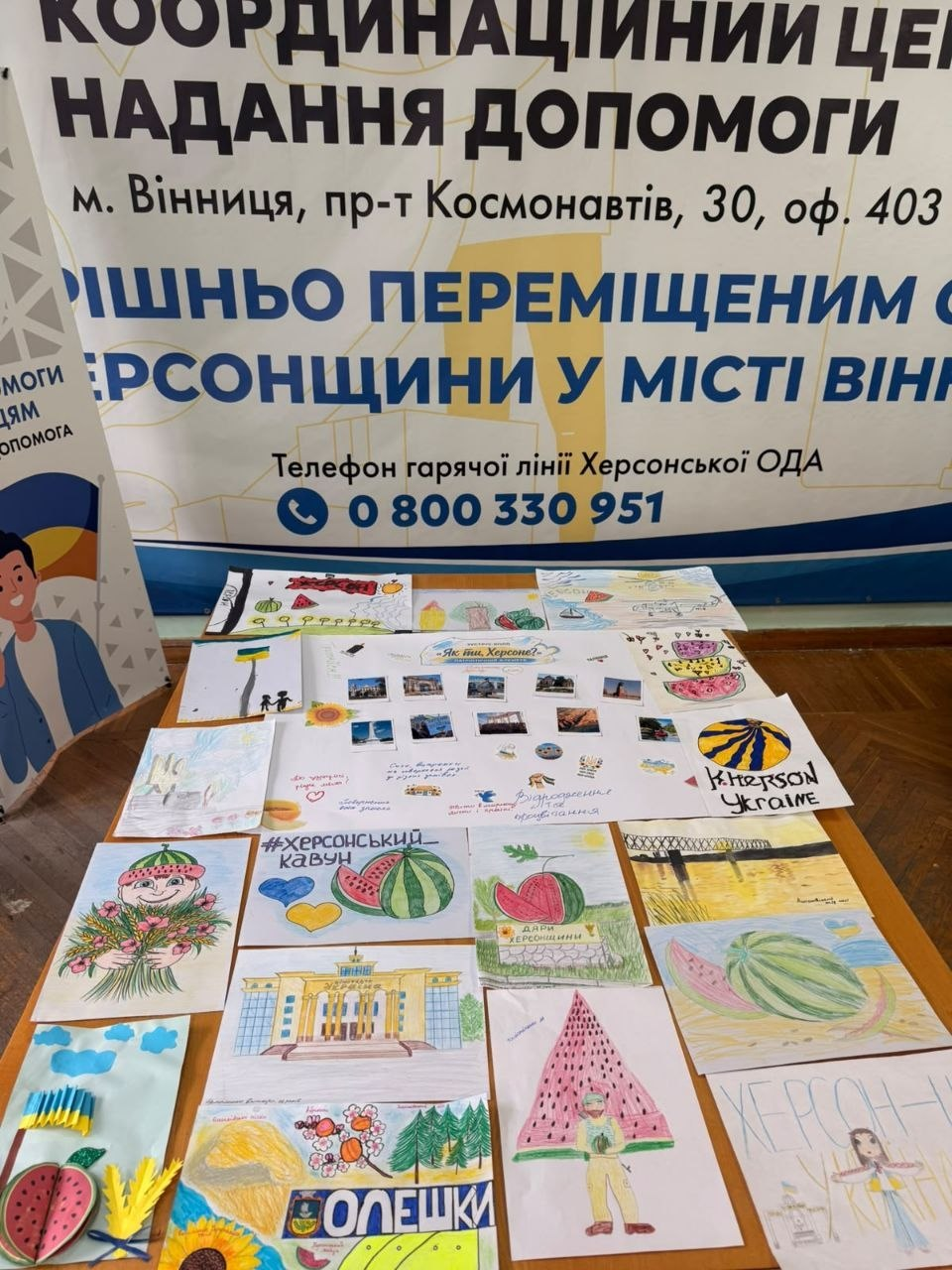 Велика виставка дитячих малюнків «Херсонщина очима дітей» розкладена на столі в приміщенні Координаційного центру «Вільні разом» у Вінниці в межах системної підтримки переселенців з Херсонщини. На передньому плані представлено творчі роботи дітей із зображенням стиглих херсонських кавунів, краєвидів Олешківських пісків із написом «ОЛЕШКИ», будівлі Херсонського художнього музею та синьо-жовтих прапорів і сердець із побажаннями. Позаду столу на дерев'яному паркеті встановлено великий інформаційний банер із написом «КООРДИНАЦІЙНИЙ ЦЕНТР НАДАННЯ ДОПОМОГИ ВНУТРІШНЬО ПЕРЕМІЩЕНИМ ОСОБАМ З ХЕРСОНЩИНИ У МІСТІ ВІННИЦЯ», де вказано номер телефону гарячої лінії Херсонської ОДА [0 800 330 951], що ілюструє патріотичну творчість маленьких херсонців в евакуації.