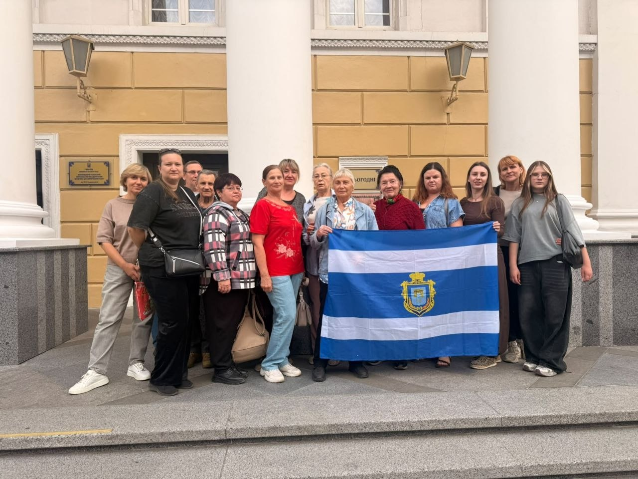 Група переселенців з Херсонщини урочисто позує для спільного фото на вулиці біля Вінницького академічного музично-драматичного театру імені М. К. Садовського до Дня міста Херсон, що відображає системну підтримку ВПО від Координаційного центру «Вільні разом» у Вінниці. Учасниці заходу вишикувалися в один ряд на кам'яному ґанку перед входом до величної будівлі з білими колонами на тлі жовто-бежевої стіни класичної архітектури. Жінки різного віку одягнені у повсякденний одяг, а по центру та праворуч вони розгорнули великий синьо-білий прапор із трьома горизонтальними смугами та золотим гербом міста Херсона, що ілюструє згуртованість громади в евакуації та організацію культурного дозвілля з відвідуванням рок-опери «Біла ворона».