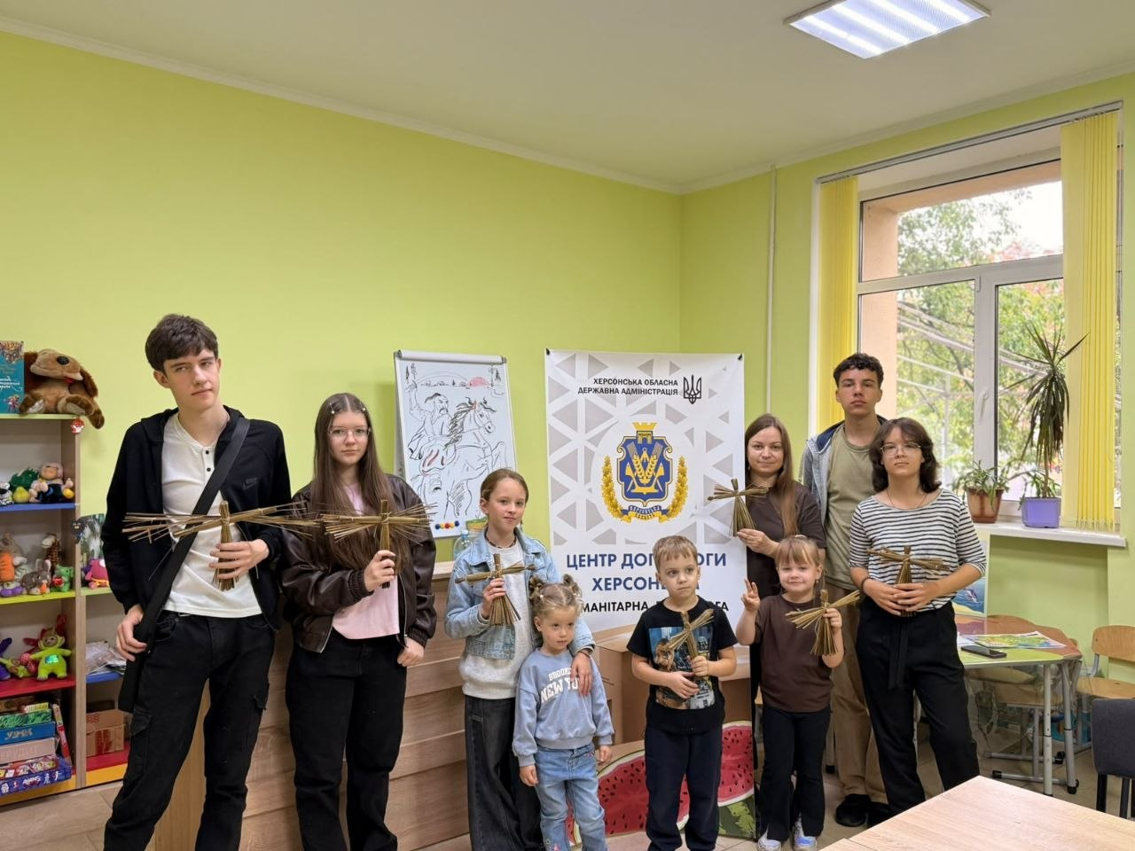 Група підлітків, дітей та дорослих урочисто позує для спільного фото у світлій кімнаті Координаційного центру «Вільні разом» у Вінниці під час екологічних ініціатив до Всесвітнього дня прибирання «World Cleanup Day 2026», організованих для системної підтримки переселенців з Херсонщини. Учасники заходу вишикувалися в один ряд по центру приміщення з ніжно-зеленими стінами, тримаючи в руках саморобні ляльки та птахи-обереги, створені під час майстер-класу з плетіння з природного рогозу після прослуховування пізнавального еколекторію від працівників Бібліотеки-філії №13 Вінницької міської ЦБС. На задньому плані за дітьми встановлено великий білий офіційний банер із тризубом, гербом області та написами «Херсонська обласна державна адміністрація» і «Центр допомоги херсонцям», ліворуч у кімнаті видно стелаж з іграшками, а праворуч — велике вікно, що відображає долучення родин переселенців до корисного творчого дозвілля.