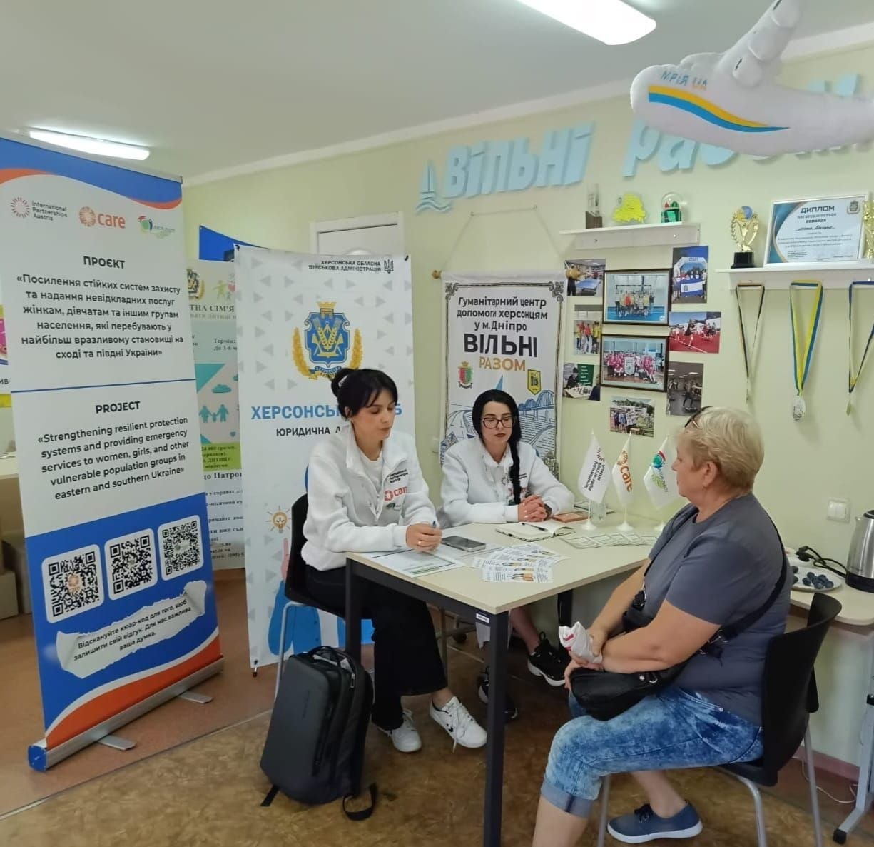 Дві фахівчині у білому фірмовому одязі приймають та консультують відвідувачку за робочим столом у кабінеті Гуманітарного центру «Вільні разом» у Дніпрі в межах надання всебічної, юридичної та соціальної допомоги переселенцям з Херсонщини. Ліворуч за столом сидить темноволоса консультантка у білому худі з емблемою організації «Care» і робить записи у документах, а поруч із нею перебуває її колега в окулярах та білому халаті, які спілкуються з жінкою у сірій футболці та синіх джинсах, що сидить перед ними на стільці. На столі розкладено інформаційні брошури та встановлено маленькі прапорці, а на задньому плані кімнати з ніжно-зеленими стінами розміщено великі стенди з написами «Херсонська обласна військова адміністрація», «Гуманітарний центр допомоги херсонцям у м. Дніпро ВІЛЬНІ РАЗОМ» та матеріали проєкту підтримки вразливих груп населення на сході та півдні України, що відображає роботу юрисконсультів благодійних фондів і громадських організацій, які загалом надали двадцять чотири консультації.