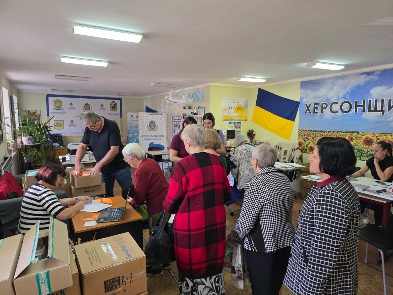 У просторому світлому залі Координаційного центру «Вільні разом» у Дніпрі велика група переселенців з Херсонщини вишикувалася в чергу біля робочих столів волонтерів під час видачі різнопланової гуманітарної допомоги, що надається в межах всебічної підтримки ВПО. Ліворуч зафіксовано працівників хабу, які опрацьовують списки та реєструють відвідувачів на ноутбуці поруч із великими картонними коробками, з яких землякам видавали рис, майонез, печиво, новий одяг, взуття, окуляри, ліки, мийні засоби та підгузки з пелюшками для маломобільних людей. На задньому плані кімнати та на стінах розміщено офіційні банери з написами «ХЕРСОНСЬКИЙ ХАБ», великий плакат «Херсонщина» із соняшниковим полем, а також висить синьо-жовтий Державний Прапор України, що ілюструє масштабну видачу допомоги від партнерських благодійних фондів та організацій.