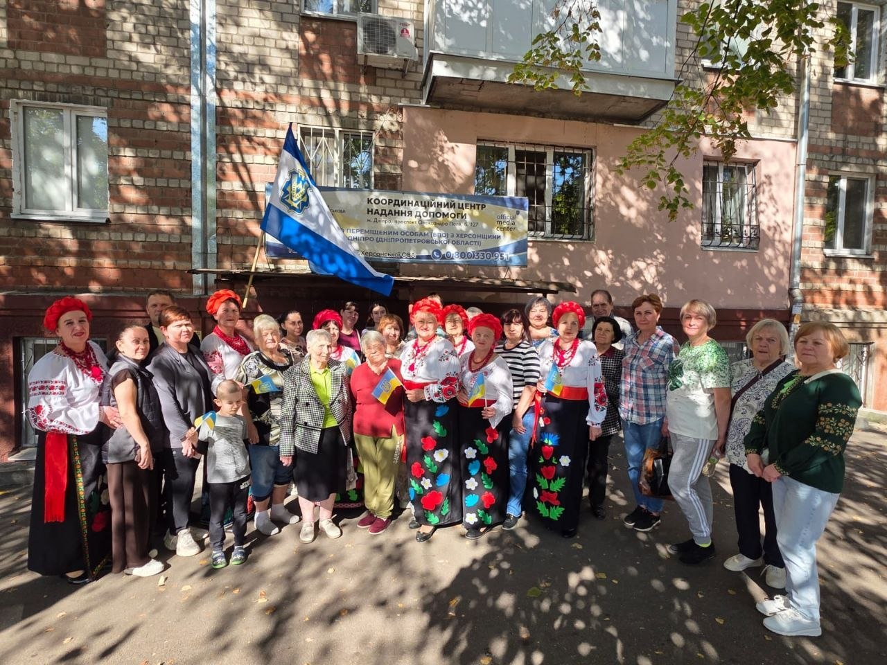 Велика група людей різного віку урочисто позує на вулиці біля входу до будівлі Координаційного центру «Вільні разом» у Дніпрі, де в межах всебічної підтримки переселенців з Херсонщини організували тематичну концертну програму до Дня міста Херсон та Дня української музики. Учасники заходу вишикувалися на асфальтованому тротуарі на тлі цегляного будинку, а серед присутніх виділяються жінки в традиційних українських вишиванках, довгих спідницях із квітковими аплікаціями та яскраво-червоних віночках. Деякі люди тримають у руках маленькі синьо-жовті прапорці України, а ліворуч над ними майорить великий прапор Херсонської області з офіційним гербом. Позаду групи на фасаді закріплено інформаційний банер центру надання допомоги, що ілюструє святкову патріотичну зустріч та згуртованість херсонської громади в евакуації.