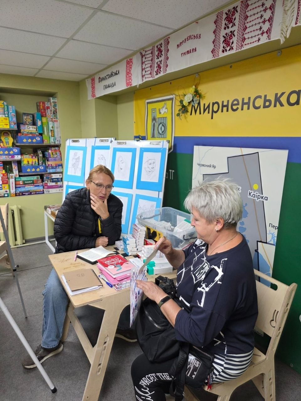 Дві жінки спілкуються за невеликим дерев'яним столом у кабінеті Координаційного центру «Вільні разом» у Дніпрі, де під час чергового заняття клубу «Будь в ресурсі» з арттерапевтичною складовою від IRC одна з них розглядає друковані матеріали в межах всебічної підтримки переселенців з Херсонщини. Ліворуч за столом сидить жінка в окулярах та чорній куртці, яка підперла підборіддя рукою й уважно слухає співрозмовницю, а праворуч на стільці перебуває сивочола відвідувачка у темній кофті, яка вивчає роздруковані аркуші. На столі перед ними лежать блокноти, ручки, брошури та книги, а на задньому плані кімнати встановлено плакат із картою Мирненської громади та великий синьо-жовтий банер із написом «Мирненська», над яким закріплено вишиті рушники з назвами територіальних громад. Ліворуч у кабінеті облаштовано білий стенд із малюнками у блакитних рамках, а в кутку видно полиці з настільними іграми, що ілюструє проведення заходів із психоемоційного розвантаження для сімей ВПО.