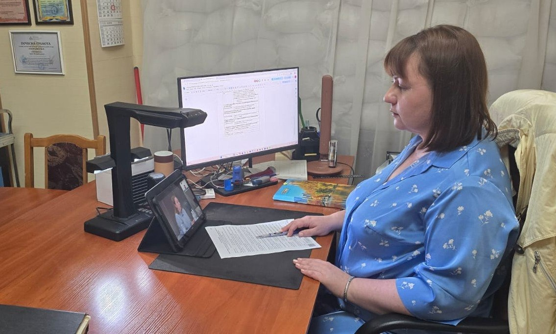 Директорка Державного архіву Херсонської області Ірина Лопушинська сидить за робочим столом під час онлайн-засідання Міжвідомчої робочої групи з координації заходів із збереження документальних свідчень про війну. Вона показана у профіль перед планшетом із відеоконференцією та монітором комп'ютера, тримаючи руку біля документів на робочому килимку. Кадр відображає участь у нараді під керівництвом Голови Державної архівної служби Анатолія Хромова щодо формування комплексного «Архіву Війни за Незалежність», документування досвіду ВПО та фіксації результатів, зокрема створення в Держархіві Херсонської області чотирьох тематичних фондів.