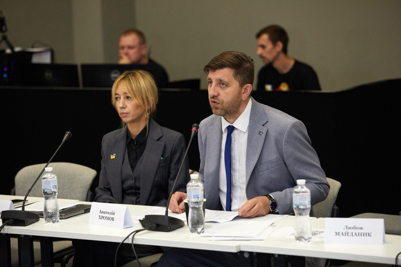 Голова Державної архівної служби України Анатолій Хромов у сірому піджаку та синій краватці виступає перед мікрофоном, очолюючи засідання Міжвідомчої робочої групи з питань збереження документальних свідчень про війну. Він сидить за столом із документами, водою та іменною табличкою, а поруч розмістилася колега у темному діловому костюмі-трійці з патріотичним значком. Кадр відображає обговорення формування комплексного «Архіву Війни за Незалежність», збереження доказової бази та обліку профільних фондів, зокрема у Державному архіві Херсонської області.