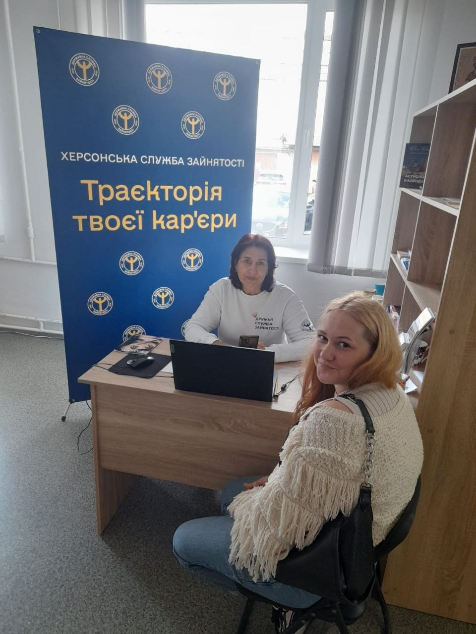 ахівчиня Бериславської філії Херсонського обласного центру зайнятості консультує дівчину щодо працевлаштування за робочим столом у кабінеті Координаційного центру «Вільні разом» у Житомирі. Зустріч проходить у межах системної та всебічної підтримки переселенців з Херсонщини. Спеціалістка у білому брендованому світшоті сидить перед ноутбуком, а поруч на стільці перебуває молода світловолоса відвідувачка у світлому в'язаному светрі та джинсах. Кадр зроблено на тлі великого синього офіційного банера з круглими емблемами установи та написами «ХЕРСОНСЬКА СЛУЖБА ЗАЙНЯТОСТІ» і «Траєкторія твоєї кар'єри». Праворуч у кімнаті біля великого вікна розташовано дерев'яний стелаж із брошурами та книгами.