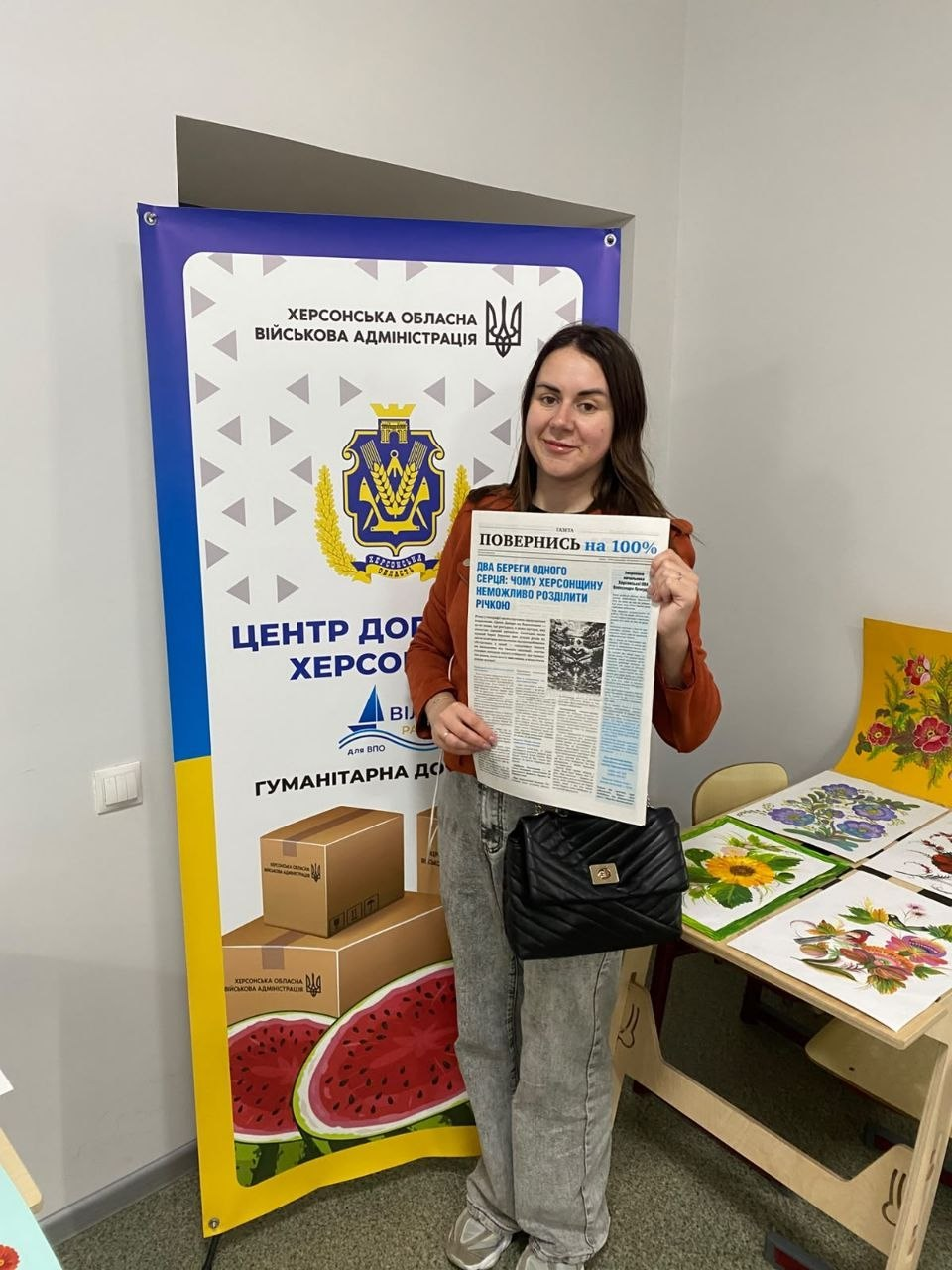 Молода жінка у коричневій кофті та сірих штанах стоїть у кабінеті Координаційного центру «Вільні разом» у Житомирі та демонструє розгорнуту друковану газету із заголовком «ПОВЕРНИСЬ на 100%» на тлі великого офіційного банера. Світлина ілюструє всебічну підтримку та інформування переселенців з Херсонщини, яким протягом тижня видавали примірники видання в центрі, а також передали їх до Коростенської та Звягельської громад. Ліворуч встановлено вертикальний банер «Херсонська обласна військова адміністрація» з гербом області та написом «ЦЕНТР ДОПОМОГИ ХЕРСОНЦЯМ», а праворуч на столі розкладено барвисті дитячі малюнки із соняшниками.