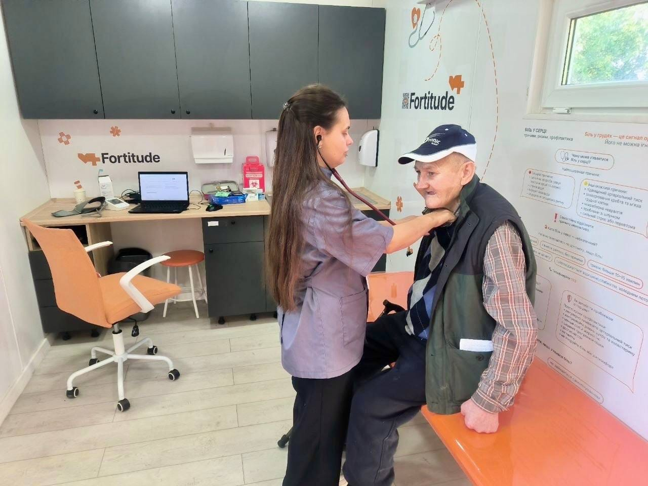 Медична працівниця медичної команди Fortitude за допомогою стетоскопа прослуховує легені літнього чоловіка у спеціально облаштованому кабінеті під час виїзного прийому, організованого для надання безкоштовної медичної допомоги жителям Великоолександрівської, Високопільської та Кочубеївської громад. Лікарка з довгим волоссям стоїть по центру кадру, а літній пацієнт у темній безрукавці та синій кепці сидить на медичній кушетці. На задньому плані облаштовано сучасну робочу зону з ноутбуком та медичним обладнанням під навісними шафами, а на білій стіні кабінету розміщено офіційні логотипи «Fortitude» та інформаційні плакати, що відображає роботу бригади лікарів, які провели обстеження й прийняли понад 480 пацієнтів у дев'яти населених пунктах Херсонщини.