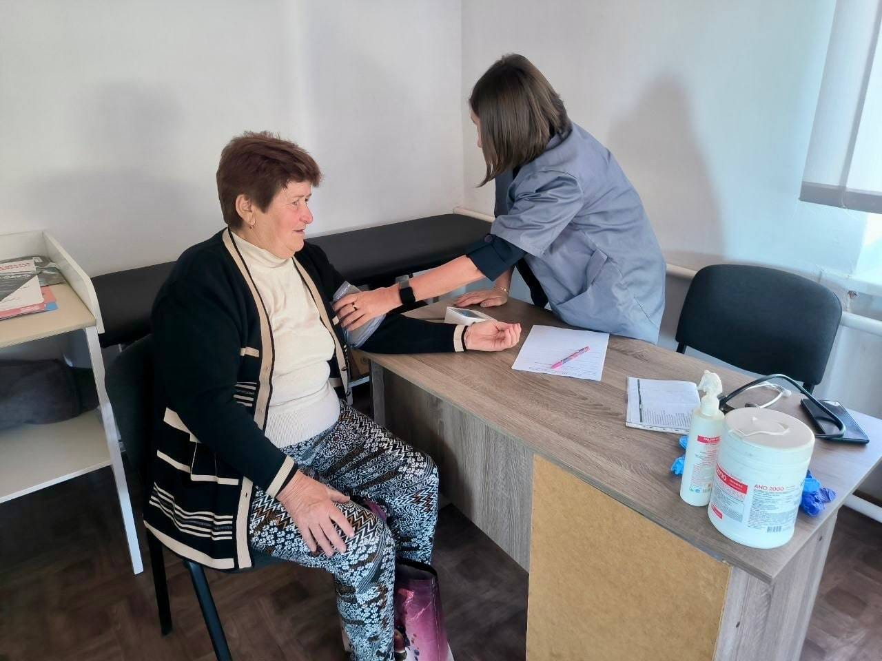 Медична працівниця у сірому халаті вимірює артеріальний тиск літній жінці за дерев'яним столом у світлому кабінеті під час виїзного прийому медичної команди Fortitude, яка протягом минулого тижня надавала безкоштовну медичну допомогу жителям Великоолександрівської, Високопільської та Кочубеївської громад. Ліворуч на стільці сидить літня відвідувачка у чорній кофті та візерунчастих штанях, а праворуч за столом схилилася лікарка, яка закріплює манжету електронного тонометра на її руці. На столі перед ними розкладено бланки, блокнот, а також встановлено медичні дезінфікуючі засоби, що відображає проведення експрес-діагностики бригадою сімейних та профільних лікарів, які прийняли понад 480 пацієнтів у дев'яти населених пунктах Херсонщини.