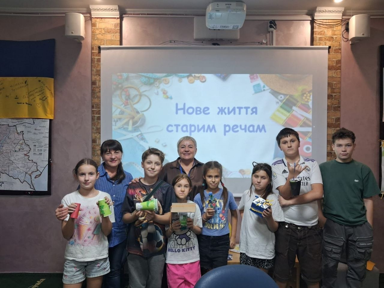 Дві дорослі жінки та семеро дітей урочисто позують у залі Координаційного центру «Вільні разом» в Ужгороді на тлі великого проєкторного екрана з презентаційною заставкою «Нове життя старим речам», демонструючи саморобні вироби з майстер-класу «Друге життя старим речам», організованого до Всесвітнього дня прибирання в межах комплексної підтримки внутрішньо переміщених осіб з Херсонщини. Учасники екологічного заходу вишикувалися в один ряд по центру кімнати: ліворуч стоїть дівчинка у білій футболці поруч із директоркою опорного ліцею у синій смугастій сорочці, а в центрі перебуває керівниця відділу освіти селищної ради у коричневій блузі з офіційним бейджем, навколо якої згрупувалися хлопчики та дівчатка, які посміхаються та тримають у руках виготовлені підставки й вироби з підручних матеріалів. Ліворуч на стіні кабінету висить карта України та підписаний синьо-жовтий Державний Прапор України, що ілюструє організацію корисного творчого й виховного дозвілля для родин херсонців в евакуації.