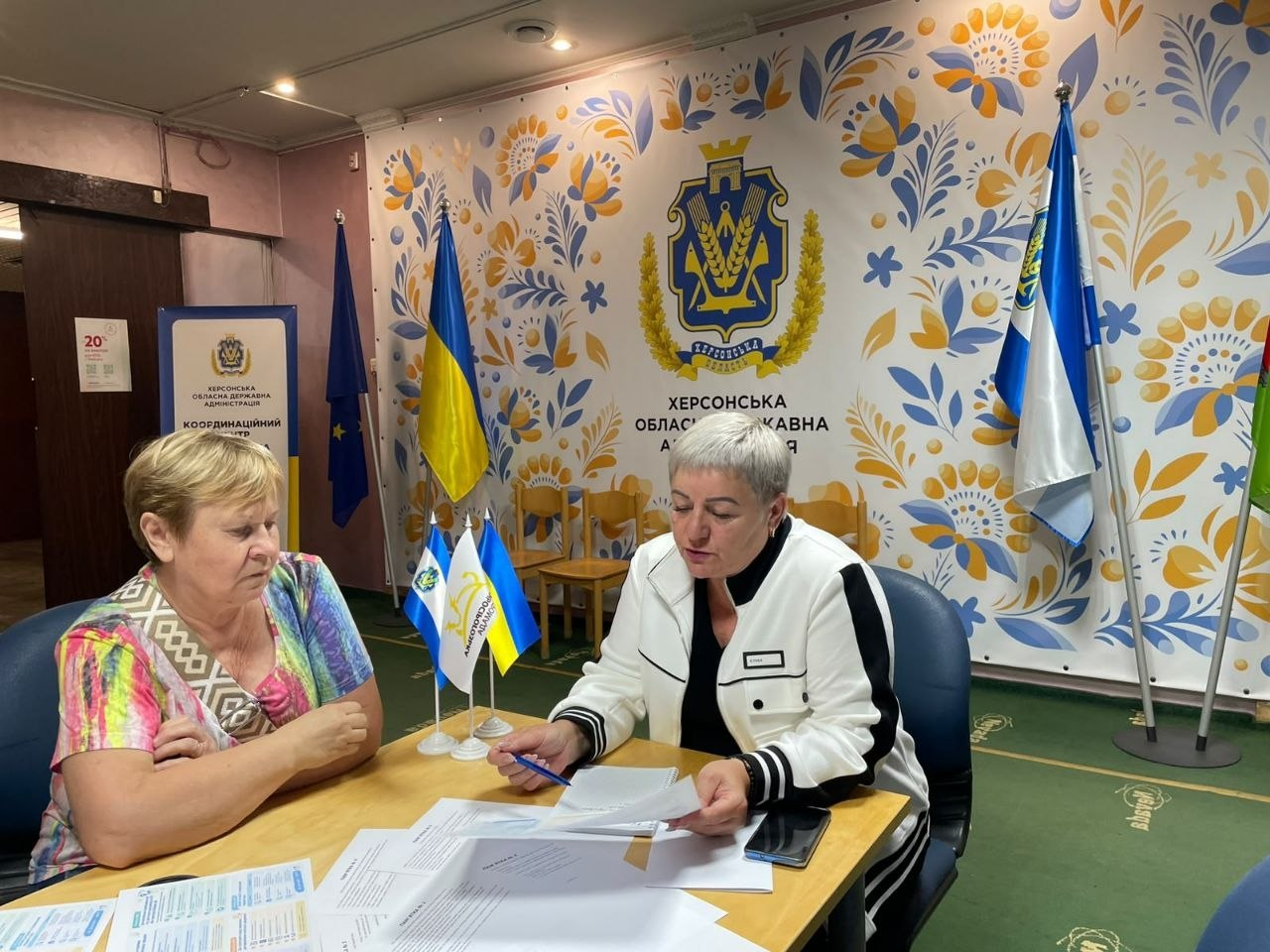 Начальниця відділу освіти, культури, молоді та спорту Нижньосірогозької селищної ради у білому жакеті консультує відвідувачку в кабінеті Координаційного центру «Вільні разом» в Ужгороді. Посадовиця робить нотатки у бланках, а навпроти неї сидить літня жінка у яскравій футболці. Кадр зроблено на тлі великого синьо-жовтого банера з гербом області та написом «ХЕРСОНСЬКА ОБЛАСНА ВІЙСЬКОВА АДМІНІСТРАЦІЯ», що ілюструє надання комплексної підтримки внутрішньо переміщеним особам з Херсонщини, проведення консультацій щодо відновлення освітніх документів «Новий старт» та підготовки до НМТ і вступу.