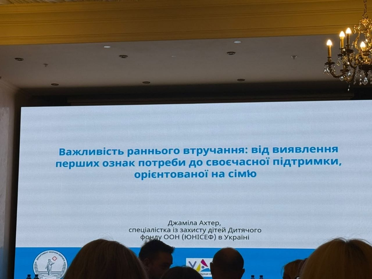 Великий мультимедійний екран у залі транслює слайд презентації спеціалістки ЮНІСЕФ Джаміли Ахтер під час Міжнародної конференції «Школа раннього втручання – 2026» в Одесі, у якій взяли участь працівники служб у справах дітей Херсонської області. На білому тлі екрана синім шрифтом виведено заголовок «Важливість раннього втручання: від виявлення перших ознак потреби до своєчасної підтримки, орієнтованої на сім'ю», а нижче вказано ім'я авторки. На передньому плані видно голови слухачів у залі, які аналізують сучасні методики підтримки сімей із дітьми раннього віку та моделі міжвідомчої взаємодії для надання комплексної соціально-медичної допомоги родинам в умовах викликів воєнного часу.