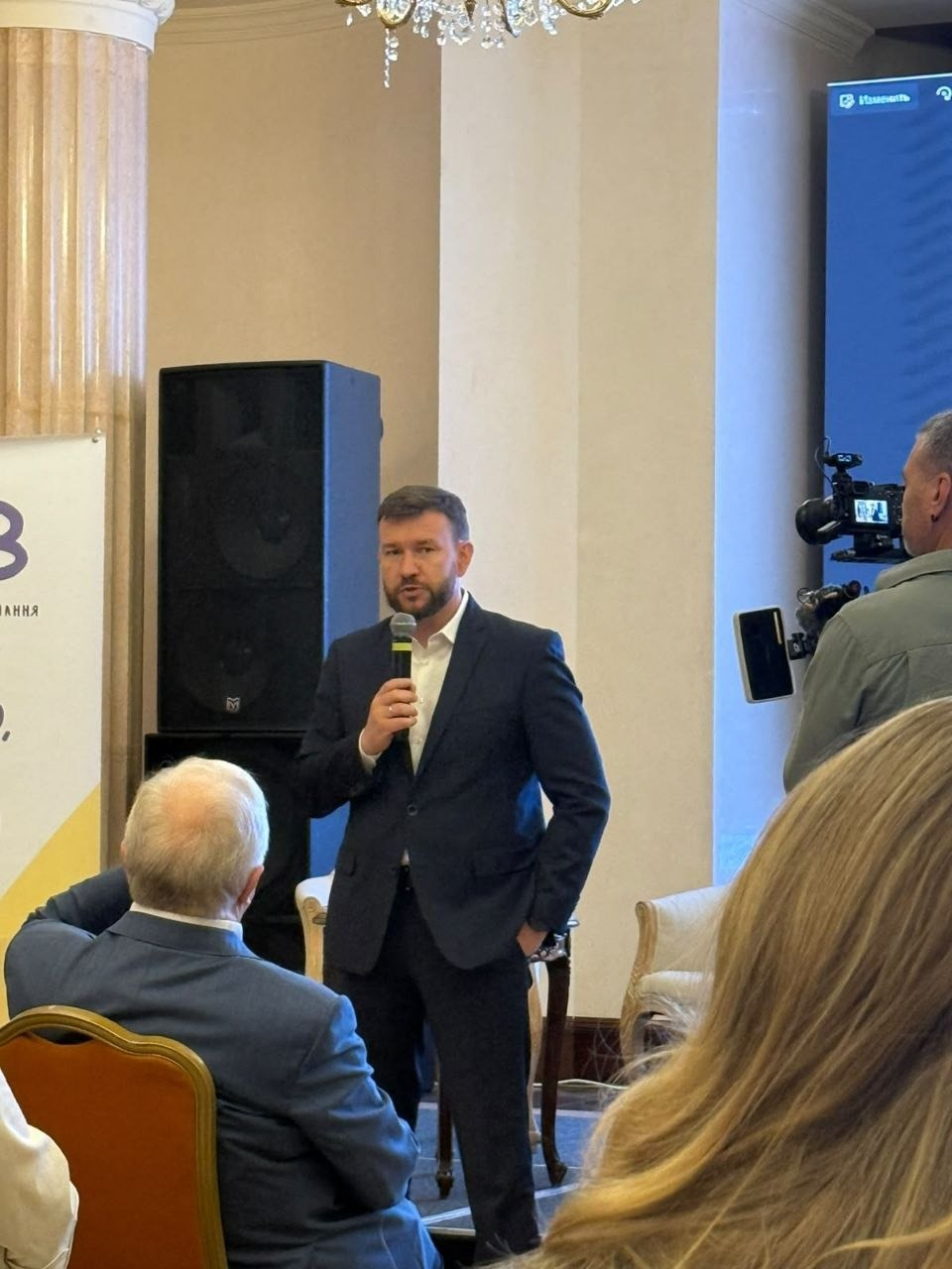 Міністр соціальної політики, сім'ї та єдності України Денис Улютін у класичному темно-синьому костюмі та білій сорочці виступає з мікрофоном на сцені під час Міжнародної конференції «Школа раннього втручання – 2026» в Одесі, до якої долучилися працівники служб у справах дітей Херсонської області. Очільник міністерства показаний по центру кадру під час промови, тримаючи ліву руку в кишені штанів. На передньому плані спиною до камери сидять слухачі, а праворуч роботу заходу знімає оператор із професійною відеокамерою. Подія, організована Центром раннього втручання КНП «Дитяча міська поліклініка №6», відображає процес обговорення сучасних методик підтримки сімей, міжвідомчої взаємодії та законодавчого закріплення послуг для надання комплексної допомоги родинам із дітьми раннього віку в умовах викликів воєнного часу.