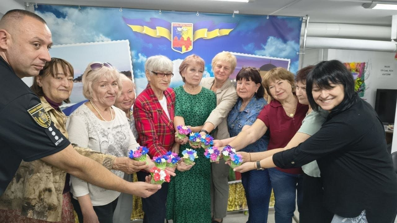 Група жінок-відвідувачок та співробітник Національної поліції України урочисто позують разом, тримаючи в руках маленькі саморобні паперові вироби, виготовлені під час творчого майстер-класу «Кошик з квітами», який провели фахівці Центру життєстійкості у Херсоні. Учасники заходу витягнули вперед руки з яскравими квітковими поробками, вишикувавшись в один ряд на тлі великого синьо-жовтого банера з гербом Херсонської області та фотографіями місцевих краєвидів. Ліворуч стоїть поліцейський у чорній формі з шевронами, а поруч із ним перебувають жінки різного віку у повсякденному одязі, які посміхаються в камеру, що ілюструє надання комплексної психосоціальної допомоги громадянам, проведення занять з арттерапії, засідань «Розмовного клубу» та консультацій щодо оформлення заяв на отримання зимової допомоги на період 2026/2027 років.