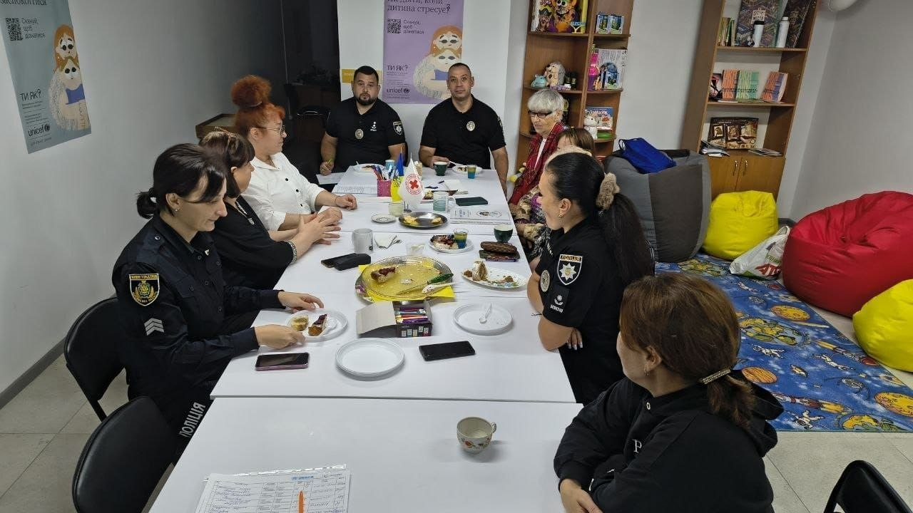 Співробітники Національної поліції України та жінки-відвідувачки за великим столом у кабінеті беруть участь у засіданні «Розмовного клубу», яке провели фахівці Центру життєстійкості у Херсоні для надання комплексної психосоціальної допомоги громадянам. Присутні розібрали алгоритми дій під час евакуації, а також державні й регіональні програми зимової підтримки на період 2026/2027 років. На білому столі перед учасниками, серед яких правоохоронці у формі, розставлено тарілки з частуваннями, чашки, канцелярію та встановлено маленькі прапорці України й Товариства Червоного Хреста України, а на стінах розміщено офіційні плакати програми ментального здоров'я «Ти як?» із логотипом ЮНІСЕФ.