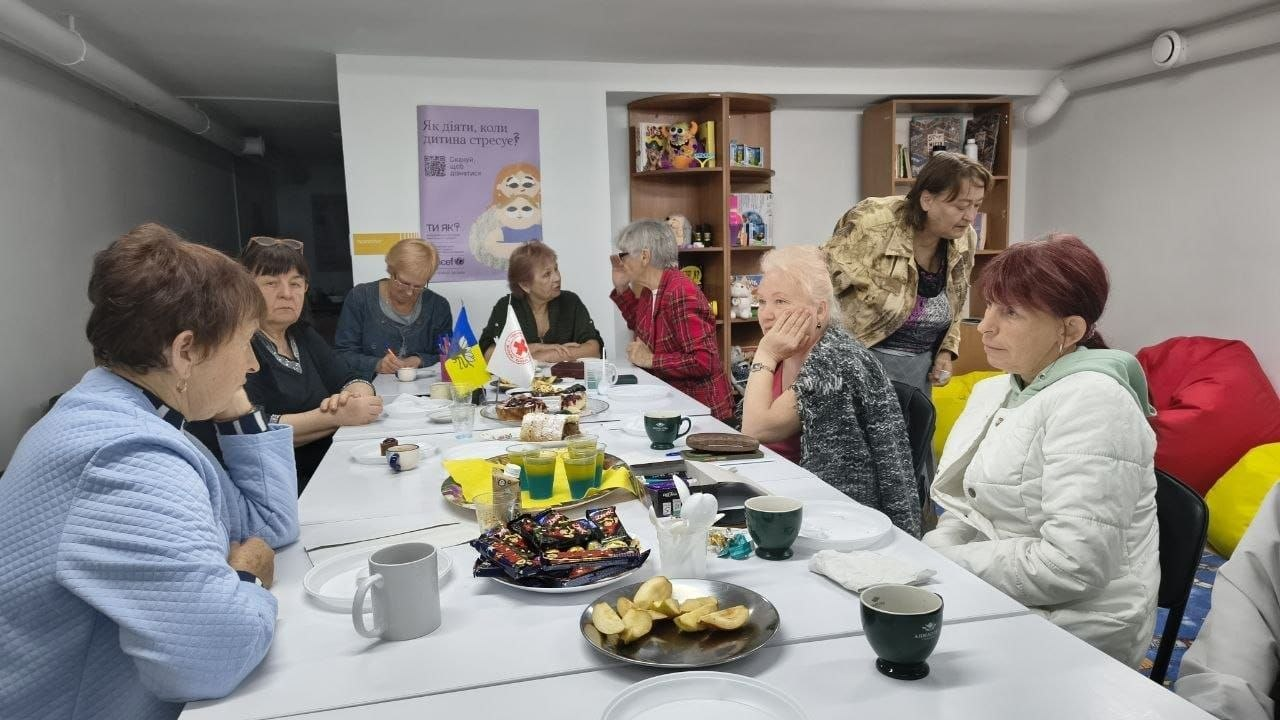 Група жінок старшого віку сидить навколо великого столу в кабінеті під час консультаційного заходу щодо коректного оформлення та подання заяв на отримання зимової допомоги, який провели фахівці Центру життєстійкості у Херсоні для надання комплексної психосоціальної допомоги громадянам у місцях тимчасового проживання. Мешканки міста зосереджено слухають інформацію, а на білому столі перед ними розставлено тарілки з частуваннями, фруктами, чашки та встановлено маленькі прапорці України й Товариства Червоного Хреста України. На задньому плані кімнати на стіні розміщено плакат програми ментального здоров'я «Ти як?» із логотипом ЮНІСЕФ, що відображає затишну та безпечну атмосферу надання необхідної підтримки людям похилого віку.