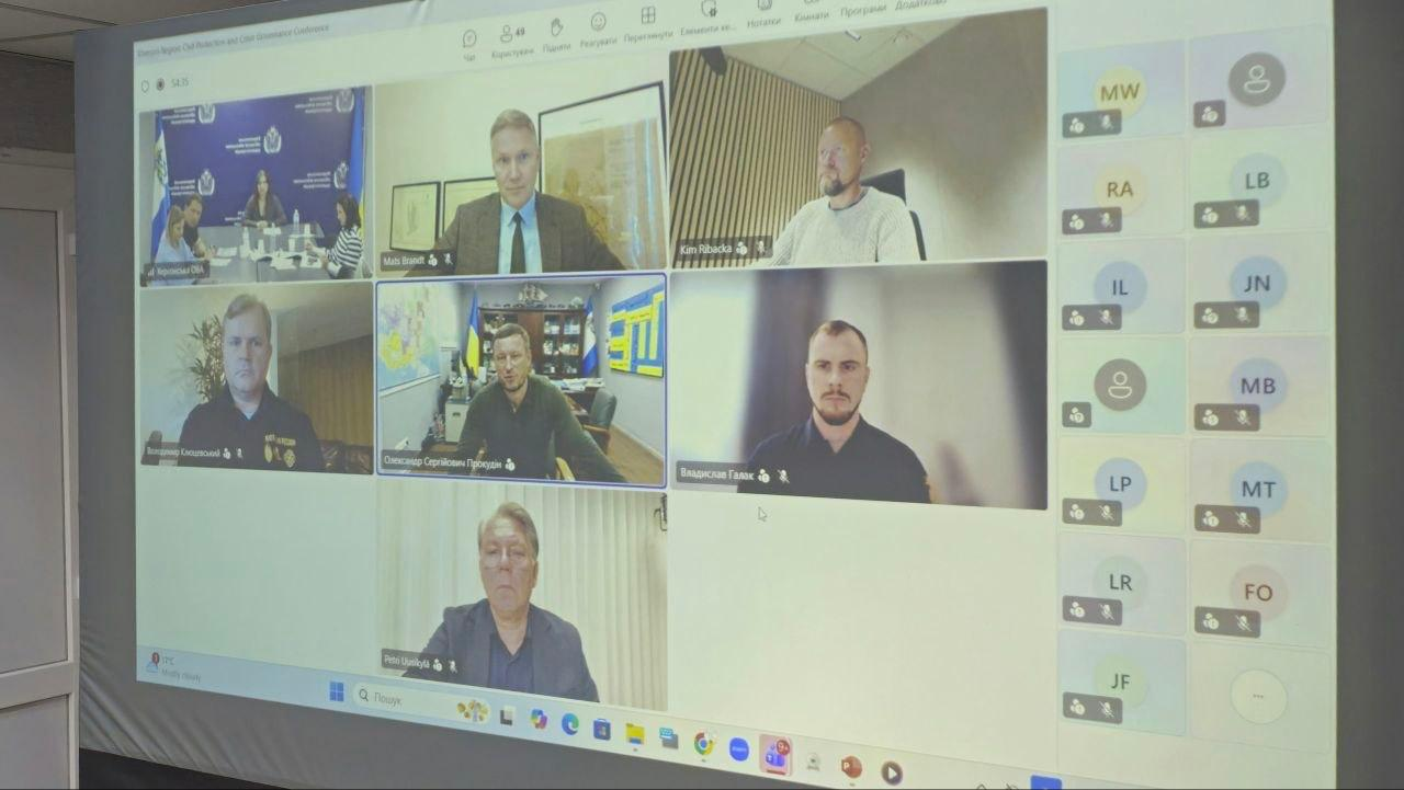 Скриншот великого проєкційного екрана демонструє спільну робочу онлайн-зустріч у форматі відеоконференції між керівництвом Херсонської ОВА та представниками фінського регіону Остроботнія, присвячену практичним рішенням для посилення стійкості громад до кризових викликів. На екрані відображається сітка з віконцями головних спікерів, де у центрі зафіксований начальник Херсонської ОВА Олександр Прокудін у робочому кабінеті, ліворуч — заступник голови Херсонської ОДА Володимир Клюцевський, а у верхньому лівому кутку — загальний вигляд зали засідань із Марією Богодуховою, Євгеном Пісоцьким та Оленою Даниловою за чорним столом. Інші віконця займають фінські партнери та українські посадовці, серед яких Mats Brandt, Ken Backman, Владислав Галак та Petro Matiytsiv, що ілюструє процес міжнародного діалогу, презентацію механізмів евакуації населення, функціонування аварійно-рятувальних служб та відновлення освітнього процесу в прифронтовому регіоні.