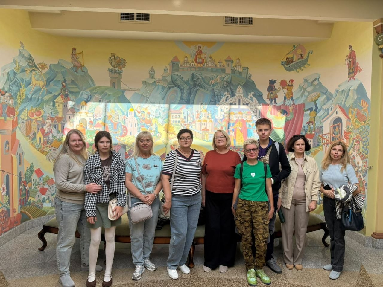 Група жінок та підлітків із числа внутрішньо переміщених осіб з Херсонщини урочисто позує в один ряд у холі будівлі на тлі яскравого панорамного настінного розпису в історично-казковому стилі в межах системної підтримки переселенців, яку надає Координаційний центр «Вільні разом» у Києві. Присутні одягнені у повсякденний одяг — джинси, кофти, футболки, дехто тримає сумки та рюкзаки. На масштабній мальовничій фресці за ними детально зображено стародавні білокам'яні фортеці, храми із золотими банями, козацькі мотиви та сюжетні сцени, що відображає процес соціокультурної інтеграції родин херсонців, організації пізнавального дозвілля та психоемоційного розвантаження в евакуації.