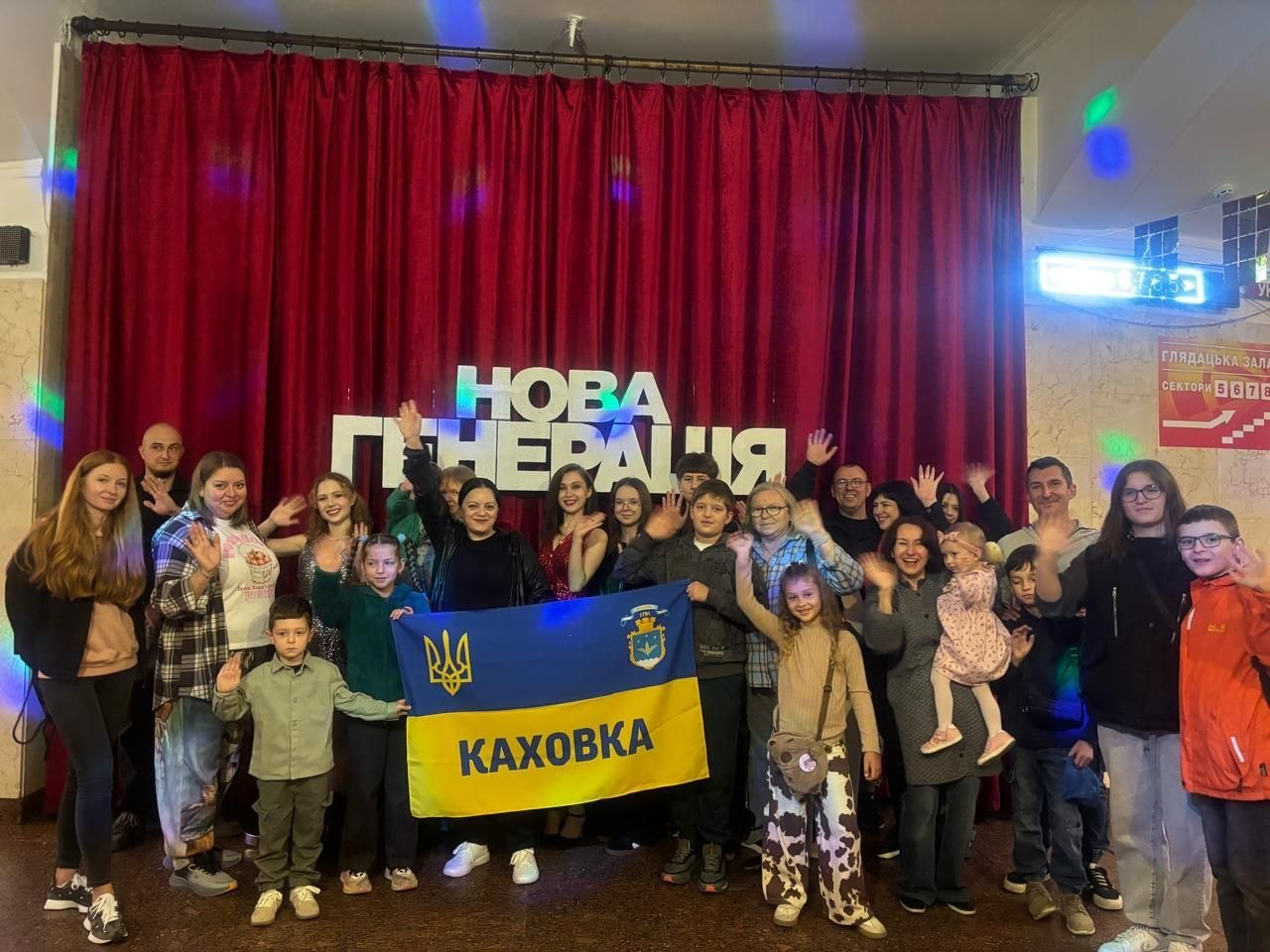 Велика група дітей та дорослих із числа переселенців із Херсонщини урочисто позує для спільного фото на тлі театральної завіси з написом «НОВА ГЕНЕРАЦІЯ» після культурно-дозвільного заходу, організованого в межах системної підтримки Координаційного центру «Вільні разом» у Києві. Учасники зустрічі посміхаються та махають руками в камеру, а по центру кадру тримають розгорнутий жовто-блакитний прапор міста Каховка з Малим Державним Гербом України та офіційним гербом міста. Світлина відображає проведення соціокультурних заходів, кінопереглядів та психоемоційного розвантаження під керівництвом фахівців хабу, що допомагає згуртуванню та підтримці каховчан та інших земляків у громаді в евакуації.