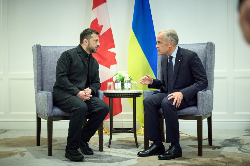 Президент України Володимир Зеленський та Прем'єр-міністр Канади Марк Карні сидять у великих сірих м'яких кріслах один навпроти одного, ведучи зосереджені переговори віч-на-віч під час офіційної зустрічі в Калгарі, де було домовлено про оборонний та бізнес-пакет допомоги на суму понад 2 мільярди доларів. Глава української держави зображений ліворуч у чорному повсякденному піджаку, штанях та темній сорочці, зчепивши пальці рук перед собою та уважно слухаючи співрозмовника. Канадський прем'єр сидить праворуч у класичному темно-синьому діловому костюмі з краваткою, нахилившись уперед і жестикулюючи правою рукою під час обговорення. Між ними на тлі встановлено вертикальні прапори Канади та України, а перед стягами стоїть невеликий круглий столик із пляшками мінеральної води та квітами, що ілюструє процес предметного діалогу щодо закупівлі ракет для Patriot, копродукції дронів, енергетичної підтримки перед зимою та участі в антибалістичній програмі FREYJA.