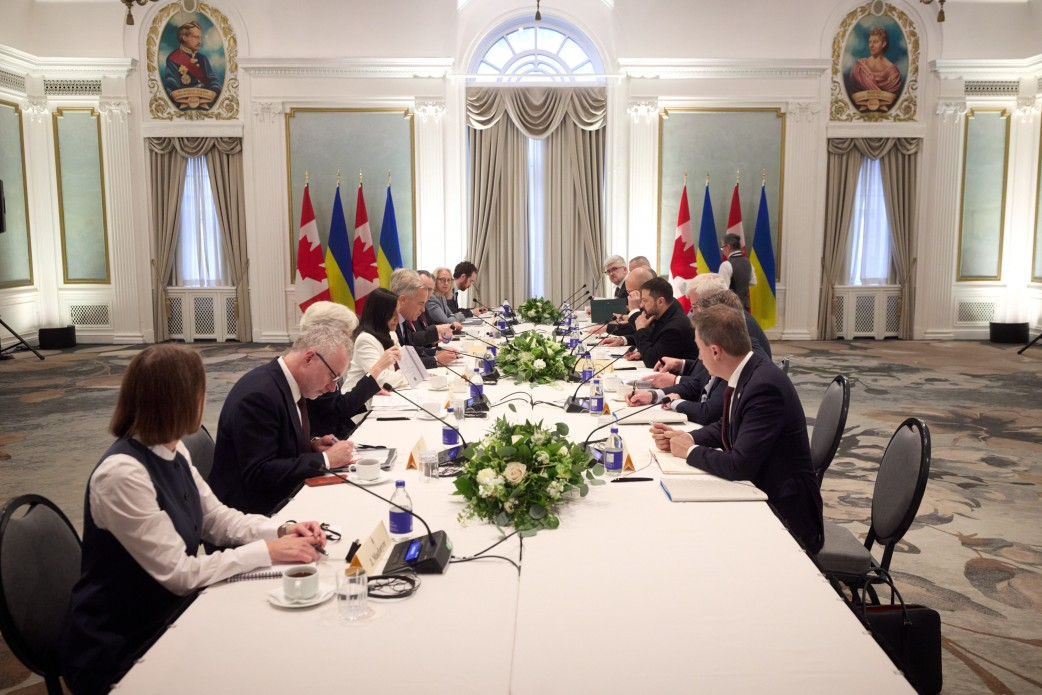 Українська та канадська делегації на чолі з Президентом України Володимиром Зеленським та Прем'єр-міністром Канади Марком Карні проводять офіційні переговори в розширеному форматі за довгим столом у залі засідань, де було досягнуто домовленостей на понад 2 мільярди доларів. Праворуч Володимир Зеленський разом із членами української команди зосереджено обговорює безпекові питання з канадськими партнерами, які сидять навпроти. Великий стіл прикрашений білими квітковими композиціями, а на задньому плані класичної світлої зали з колонами симетрично встановлено державні прапори Канади та України, що відображає процес масштабного міждержавного діалогу щодо постачання ракет для Patriot, амуніції для F-16, копродукції дронів та енергетичної підтримки перед зимою.