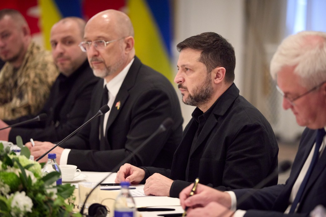 Президент України Володимир Зеленський разом із Прем'єр-міністром Денисом Шмигалем та Міністром оборони Рустемом Умєровим беруть участь в офіційних переговорах у розширеному форматі з канадською делегацією в Калгарі, де було домовлено про пакет допомоги на суму понад 2 мільярди доларів. Глава держави у чорному повсякденному піджаку сидить за довгим столом біля настільного мікрофона, зосереджено виступаючи перед партнерами. Поруч із ним ліворуч у класичному костюмі та окулярах сидить Денис Шмигаль, далі — Рустем Умєров та представник у камуфляжній формі, а на передньому плані справа видно канадського посадовця, який занотовує інформацію ручкою. На білому столі розставлено чашки, мінеральну воду та квіткові композиції, а на задньому плані видніються державні прапори, що відображає процес обговорення оборонної підтримки, постачання ракет для Patriot, амуніції для F-16, копродукції дронів та покриття бюджетного дефіциту України.