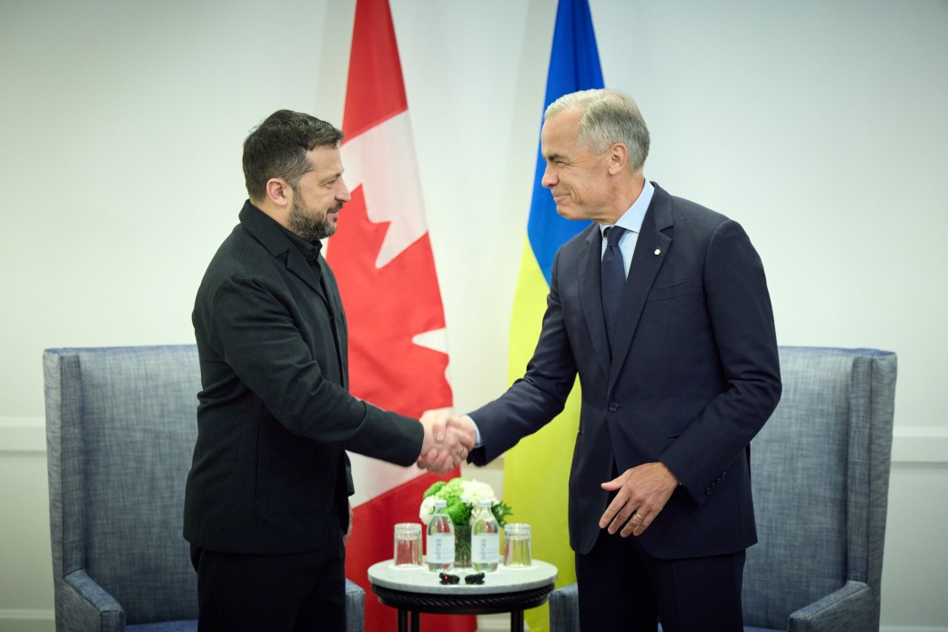 Президент України Володимир Зеленський та Прем'єр-міністр Канади Марк Карні посміхаються та тиснуть один одному руки під час офіційної зустрічі в Калгарі, де було досягнуто домовленостей на суму понад 2 мільярди доларів щодо виділення коштів на ракети для Patriot, амуніцію для F-16, копродукцію дронів та підтримку енергетики перед зимою. Глава української держави зображений ліворуч у чорному піджаку, а канадський прем'єр позує праворуч у класичному темно-синьому діловому костюмі, білій сорочці та краватці. Позаду лідерів встановлено великі вертикальні прапори: червоно-білий із кленовим листом Прапор Канади ліворуч та синьо-жовтий Державний Прапор України праворуч. Кадр ілюструє результати предметних перемовин щодо посилення протиповітряної оборони, розширення санкційного тиску, приєднання Канади до антибалістичної програми FREYJA та зміцнення всебічного безпекового партнерства між двома державами.