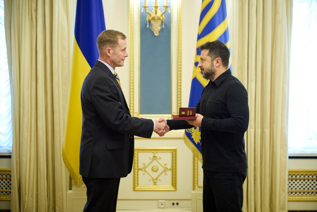 Президент України Володимир Зеленський урочисто тисне руку генерал-лейтенанту Кертісу Баззарду, вручаючи йому орден «За заслуги» III ступеня під час офіційної зустрічі в Києві. Глава держави зображений праворуч у чорній сорочці, тримаючи в руці нагородну коробочку з відзнакою, а командувач Спеціальної місії НАТО з підтримки безпеки для України стоїть ліворуч у діловому костюмі, приймаючи нагороду. Позаду них встановлено Державний Прапор України та синій прапор Президента України, що підкреслює офіційний характер зустрічі, присвяченої результатам саміту НАТО в Анкарі, розвитку програми фінансування оборонного спорядження PURL та співпраці в межах Drone Deal.