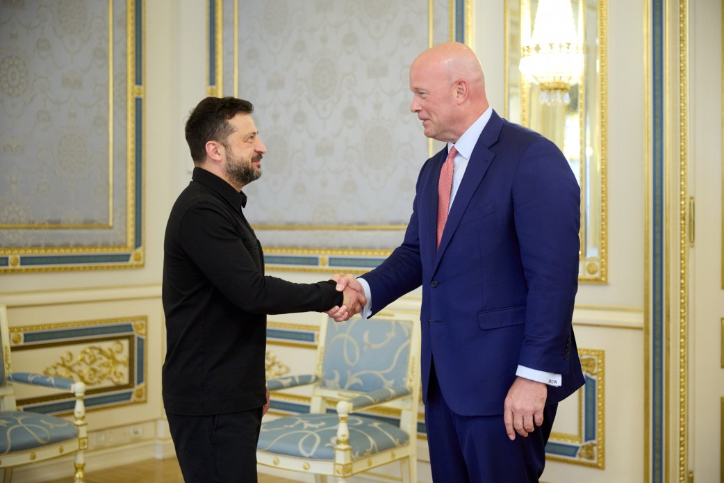 Президент України Володимир Зеленський та Посол США при НАТО Метью Вітакер посміхаються та тиснуть один одному руки під час офіційної зустрічі, де обговорили результати саміту НАТО в Анкарі, зокрема реалізацію важливих рішень для захисту українського неба та ліцензії на виробництво ракет для систем Patriot. Глава української держави зображений ліворуч у повсякденній чорній сорочці та темних штанях, а американський дипломат позує праворуч у класичному темно-синьому діловому костюмі, білій сорочці та світло-рожевій краватці. Вони стоять по центру кімнати на тлі вишуканого світлого інтер'єру з позолоченим оздобленням стін та м'якими кріслами, що ілюструє процес важливих міжнародних перемовин щодо посилення ППО, роботи над майбутньою угодою у форматі Drone Deal та розвитку успішної програми PURL із фінансування оборонного спорядження для України.