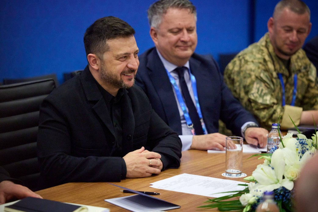 Президент України Володимир Зеленський у чорній сорочці посміхається та сидить за дерев'яним столом переговорів у конференційній залі в Анкарі разом із членами української делегації під час підписання безпекової угоди про оборонне співробітництво у форматі Drone Deal з Естонією. Глава держави зчепив пальці рук перед собою, слухаючи виступи колег, а поруч із ним за столом сидять українські посадовці, серед яких представник у класичному костюмі зі стрічкою-бейдж на шиї та військовослужбовець Сил оборони України в камуфляжній формі піксель. На столі перед ними лежать паперові документи, ручка та край великої квіткової композиції з білих лілій, що ілюструє офіційну зустріч із Прем’єр-міністром Естонії Крістеном Міхалом задля запуску спільного військового виробництва засобів ППО, дронів, сенсорних систем та боєприпасів.