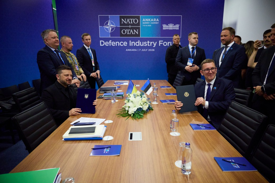 Президент України Володимир Зеленський та Прем’єр-міністр Естонії Крістен Міхал сидять один навпроти одного за великим дерев'яним столом у конференційній залі в Анкарі під час підписання двосторонньої угоди про оборонне співробітництво у форматі Drone Deal. Глава української держави зображений ліворуч у чорній сорочці, тримаючи розгорнуту синю папку з Державним Гербом України, а естонський прем'єр позує праворуч в окулярах та класичному темно-синьому костюмі з краваткою, тримаючи темну папку з гербом своєї країни. На столі між лідерами лежить композиція з білих квітів, встановлено мініатюрні настільні прапорці України та Естонії, а на задньому плані на тлі великого синього банера з логотипом НАТО та написами «Defence Industry Forum» і «ANKARA | 7 JULY 2026» стоять члени оборонних делегацій. Кадр ілюструє процес підписання вже сьомої для України угоди Drone Deal, яка посилює промислову співпрацю, обмін досвідом і технологічну копродукцію між країнами у сфері виробництва дронів, засобів ППО та боєприпасів.