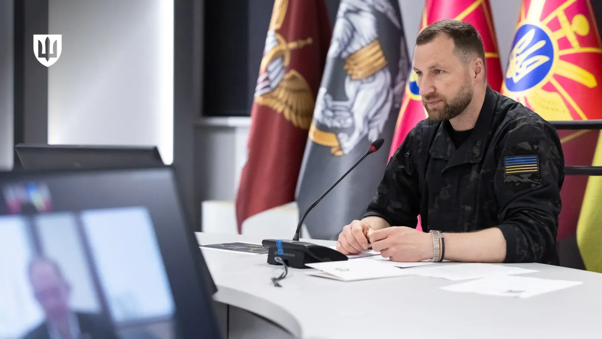 Міністр оборони України Євгеній Хмара в чорній камуфляжній формі із синьо-жовтим прапором на рукаві сидить за світлим столом у залі засідань під час телефонної розмови з Міністром оборони Данії Єппе Бруусом, присвяченої посиленню спроможностей для протидії балістичним загрозам, реактивним ударним дронам та реалізації проєкту FREYJA. Посадовець склав руки перед собою на документах біля настільного мікрофона та уважно слухає данського колегу, віконце з яким транслюється на екрані розмитого ноутбука на передньому плані ліворуч. На задньому плані за спиною очільника відомства встановлено великі офіційні прапори військових підрозділів і Збройних Сил України, а в лівому верхньому кутку кадру нанесено білий стилізований Малий Державний Герб України, що ілюструє процес обговорення критичних потреб Сил оборони, розвитку рішень у сфері перехоплювачів, радіоелектронної боротьби та поглиблення оборонної співпраці між країнами.