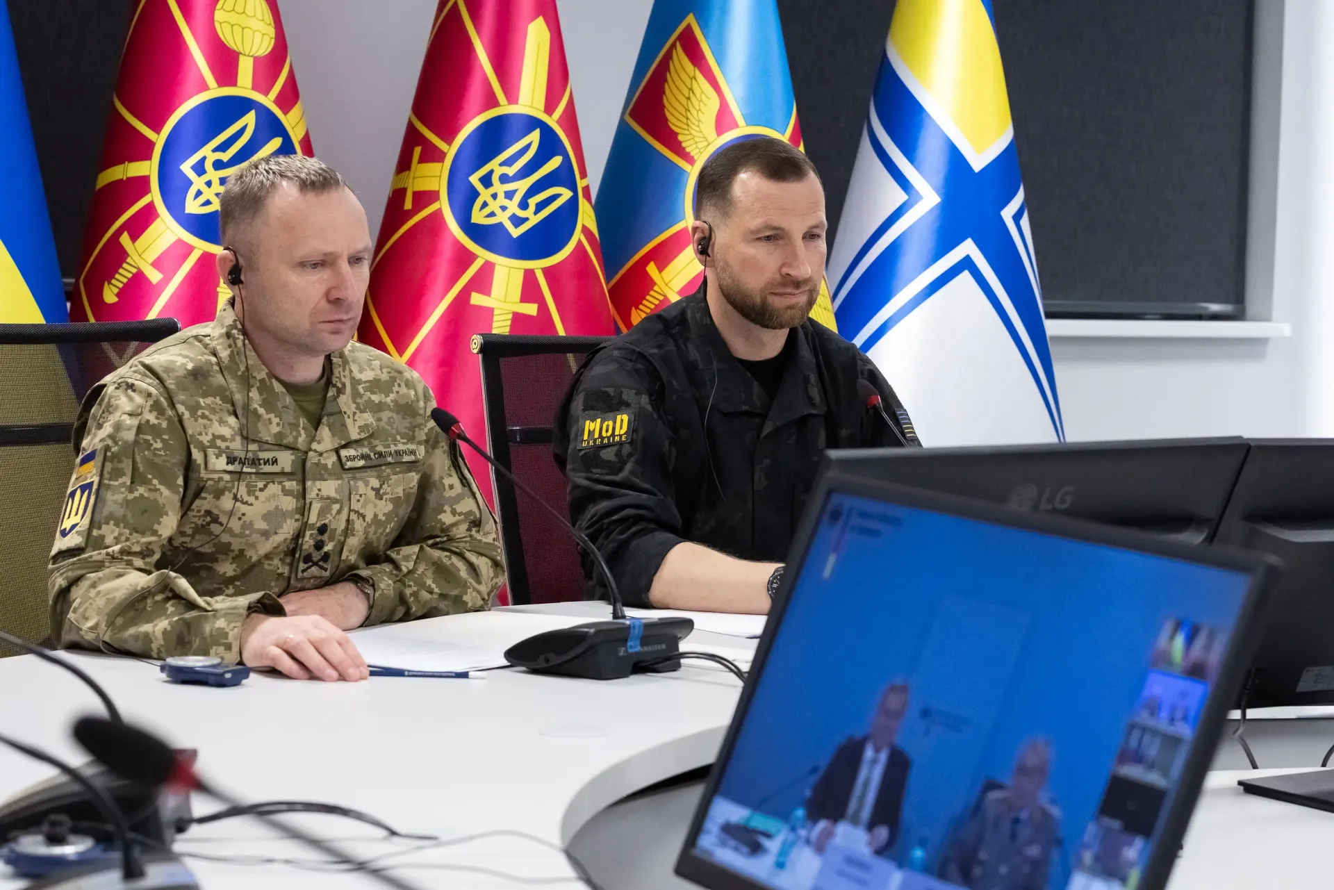 Міністр оборони України Євгеній Хмара в чорній формі з написом «MoD UKRAINE» та Головнокомандувач Збройних Сил України генерал-майор Михайло Драпатий ліворуч у камуфляжі синять поруч за світлим столом у залі засідань під час телефонної відеорозмови з Міністром оборони Данії Єппе Бруусом. Посадовці зосереджено слухають данського колегу, віконце з яким транслюється на екрані монітора на передньому плані праворуч. На задньому плані за їхніми спинами встановлено Державний Прапор України та офіційні прапори військових підрозділів і Збройних Сил України, що підкреслює офіційний характер двостороннього безпекового діалогу, присвяченого обговоренню критичних потреб Сил оборони, протидії балістичним загрозам, реактивним ударним дронам та реалізації проєкту FREYJA.