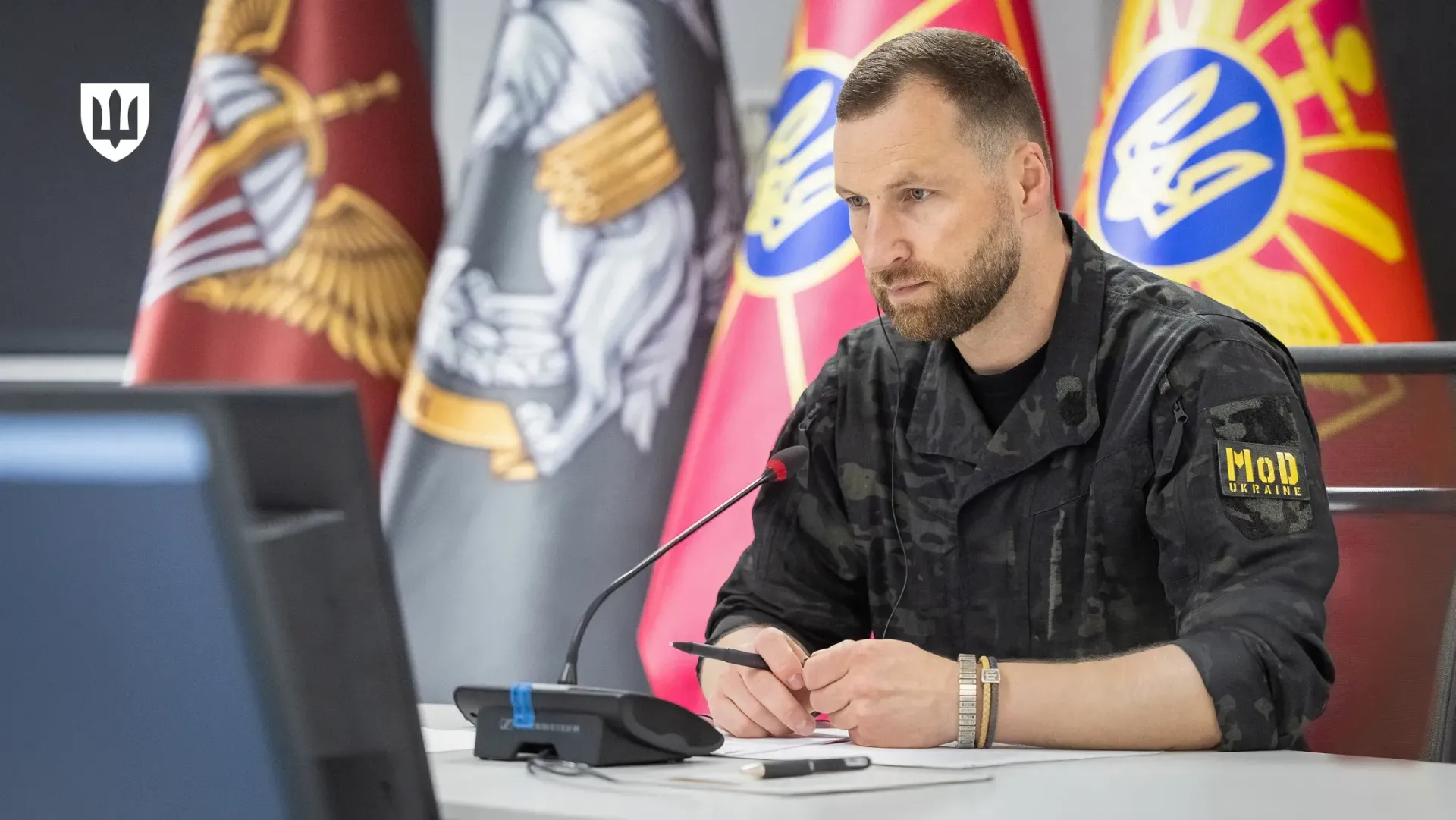 Тимчасово виконуючий обов’язки Міністра оборони України Євгеній Хмара в чорній камуфляжній формі із нашивкою «MoD UKRAINE» на лівому рукаві сидить за світлим столом у залі засідань під час телефонних розмов із Міністром оборони Швеції Полом Йонсоном та Міністром оборони Норвегії Торе Сандвіком. Посадовець зображений крупним планом по пояс, він тримає чорну ручку в руках над документами біля настільного мікрофона і зосереджено дивиться на екран монітора ліворуч, обговорюючи термінові запити від бойових підрозділів. На задньому плані за його спиною встановлено великі офіційні прапори військових підрозділів і Збройних Сил України, а в лівому верхньому кутку кадру нанесено білий стилізований Малий Державний Герб України, що відображає процес двостороннього оборонного діалогу щодо фінансування українських дронів, посилення ППО проти балістики та розвитку європейського проєкту FREYJA.