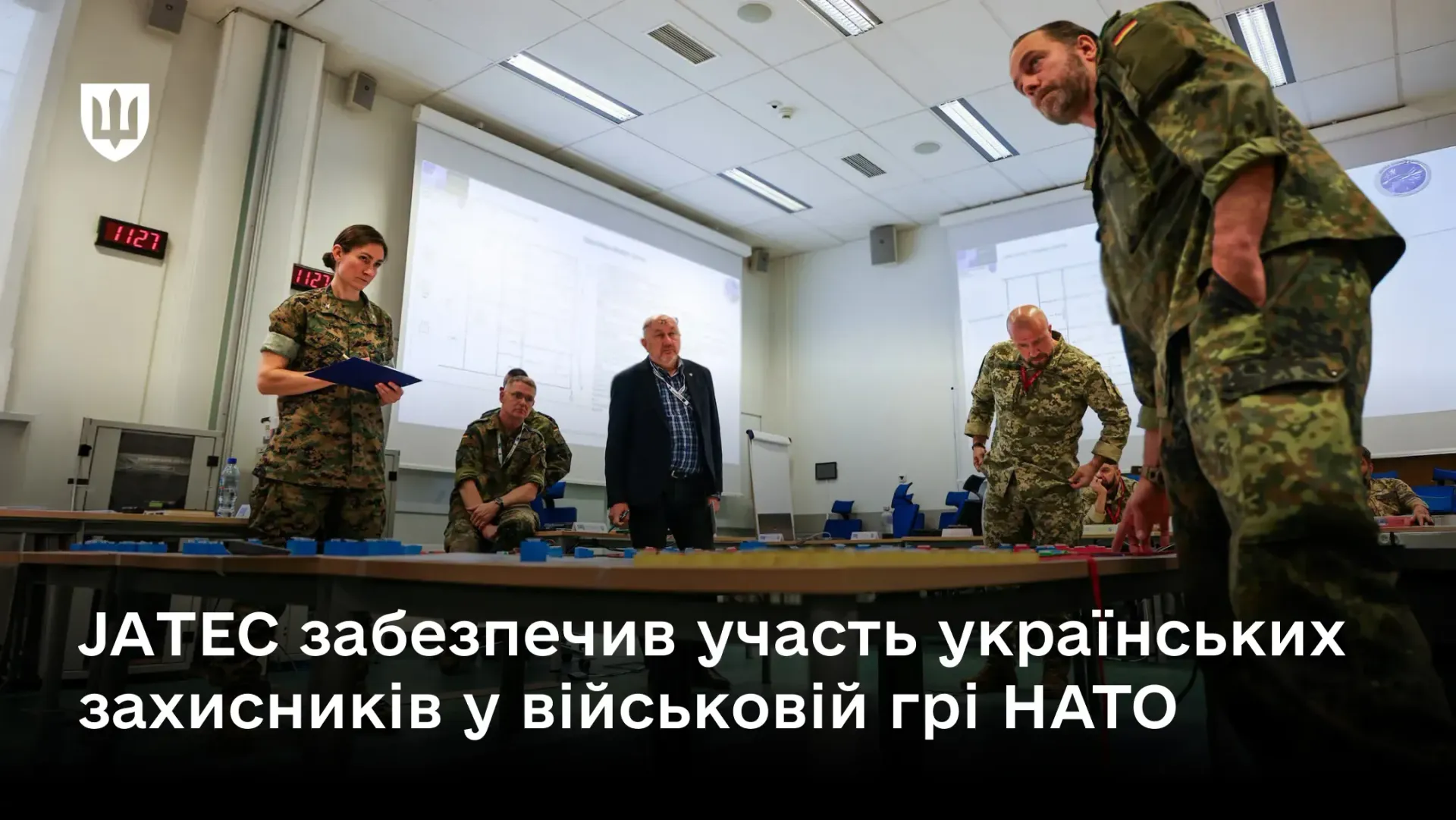 Офіційний інформаційний банер із заголовком про участь Збройних Сил України у заходах Альянсу, на якому зафіксовано процес планування операцій у межах спільної військової гри «Vistula Vanguard 26». У великій аудиторії з проєкційними екранами група військовослужбовців у камуфляжній формі країн НАТО та України зібралася навколо столів із масштабним макетом місцевості, обговорюючи тактичні процедури. На передньому плані праворуч нахилився офіцер у німецькому камуфляжі, ліворуч стоїть жінка-військовослужбовець із планшетом, а по центру перебуває цивільний інструктор. У лівому верхньому кутку розміщено білий Малий Державний Герб України, а внизу нанесено заголовок «JATEC забезпечив участь українських захисників у військовій грі НАТО», що ілюструє залучення українського бойового досвіду у сфері БпЛА, розвідки та РЕБ для підвищення реалістичності командно-штабних сценаріїв Альянсу.