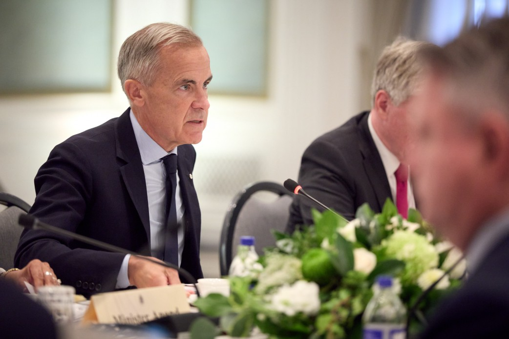 Прем'єр-міністр Канади Марк Карні бере участь в офіційних переговорах у розширеному форматі з українською делегацією в Калгарі, де було домовлено про виділення пакета допомоги Україні на суму понад 2 мільярди доларів. Канадський лідер зображений ліворуч у класичному темно-синьому діловому костюмі, білій сорочці та темній краватці, він сидить за довгим столом біля настільного мікрофона і зосереджено виступає перед учасниками зустрічі. Поруч із ним на дальньому плані сидить інший член канадської делегації у костюмі та сорочці з яскравою краваткою, а на передньому плані справа розмито видно представника української сторони. Білий стіл засідань прикрашений великою композицією з білих квітів та зелені, на ньому розставлено пляшки з водою та посуд, що відображає процес обговорення оборонної підтримки, постачання ракет для Patriot, амуніції для F-16, копродукції дронів та залучення фінансування для покриття дефіциту українського бюджету.