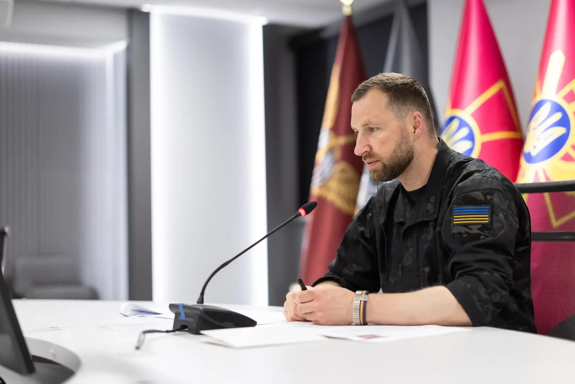 Міністр оборони України Євгеній Хмара в чорній камуфляжній формі із синьо-жовтим прапором на рукаві сидить за світлим столом у залі засідань під час телефонної розмови з Генеральним секретарем НАТО Марком Рютте, присвяченої підготовці до засідання Контактної групи у форматі «Рамштайн». Посадовець склав руки перед собою поруч із настільним мікрофоном та уважно дивиться вбік, аналізуючи першочергові потреби держави. На передньому плані ліворуч розмито видно відкритий монітор комп'ютера, а на задньому плані за спиною керівника відомства встановлено великі офіційні прапори Міністерства оборони та Збройних Сил України. Світлина ілюструє процес важливих безпекових перемовин щодо термінового постачання засобів протиповітряної оборони, залучення додаткового фінансування для Сил оборони та координації постачання критично важливого озброєння в межах програми Prioritized Ukraine Requirements List (PURL).