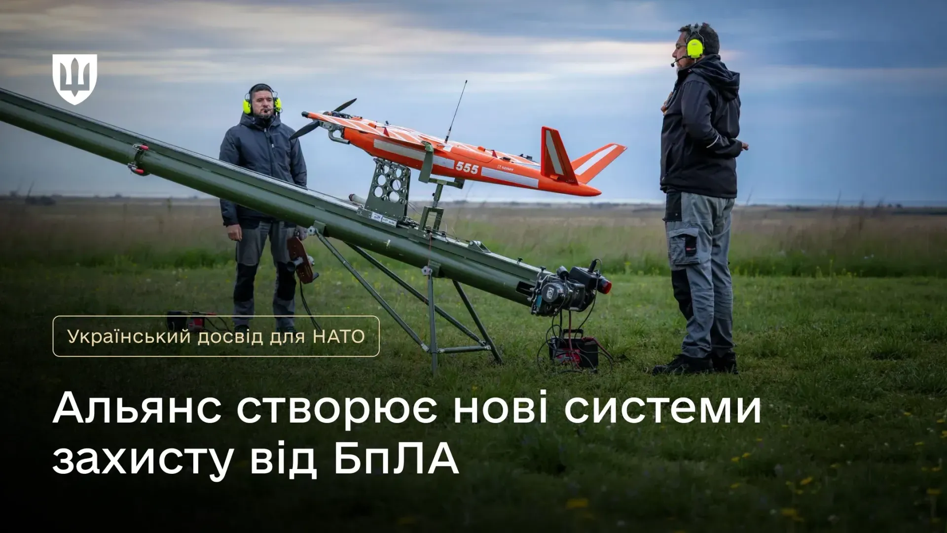 Офіційний інформаційний банер із заголовком про оборонні технології, на якому зображено підготовку до запуску безпілотника з мобільної катапульти в межах протидронової ініціативи НАТО «LCI-X». На передньому плані встановлено довгу зелену рейку пускової установки з яскраво-помаранчевим БпЛА літакового типу під номером «555», біля якого на тлі поля стоять двоє операторів у захисних навушниках. У лівому верхньому кутку розміщено Малий Державний Герб України, поруч нанесено написи «Український досвід для НАТО» та «Альянс створює нові системи захисту від БпЛА», що ілюструє залучення бойового досвіду українських експертів JATEC для розробки реалістичних сценаріїв та вдосконалення технологій перехоплення дронів Альянсом.