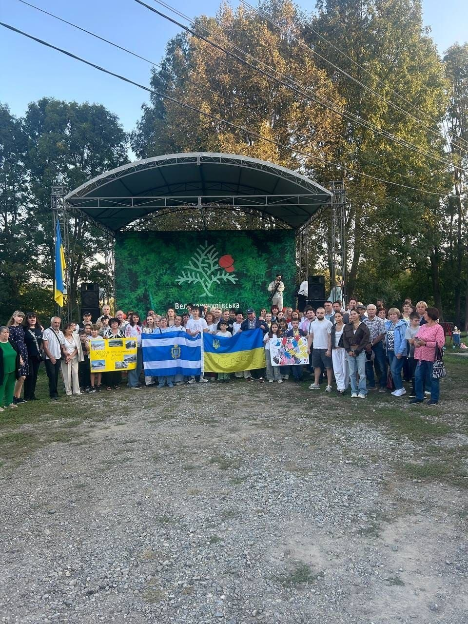Велика група дорослих і дітей із числа переселенців із Херсонщини стоїть на вулиці перед критою металевою сценою в селі Великий Кучурів під час святкування Дня Херсона та відвідування фестивалю до Дня української музики «Від Херсона до Буковини — рецепт єдності». Близько сотні херсонців зібралися на ґрунтовій галявині, тримаючи попереду Державний Прапор України, синьо-білий прапор міста Херсона з його гербом, жовтий інформаційний плакат та дитячий малюнок із різнокольоровими відбитками долонь. Задня стіна сцени прикрашена великим зеленим банером із символікою Великокучурівської громади, що ілюструє спільний патріотичний захід Координаційного центру «Вільні разом» у Чернівцях та Палацу молоді і студентів Херсонської обласної ради, спрямований на культурну інтеграцію та єднання українців в евакуації.