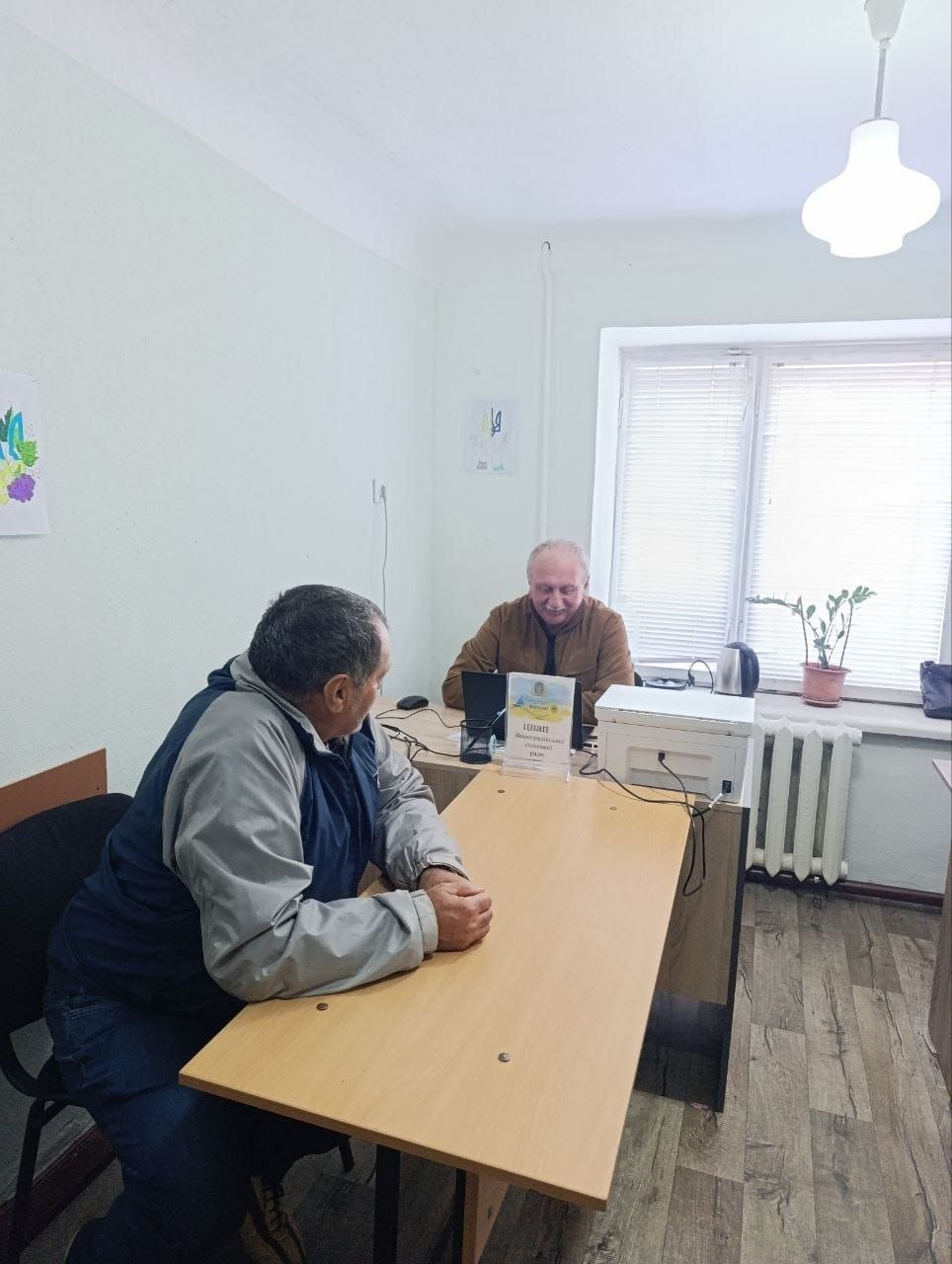 Адміністратор відділу ЦНАП Нововоронцовської селищної ради В.І. Чебан у коричневій куртці сидить за робочим столом у кабінеті в Чернівцях та здійснює прийом літнього чоловіка з нагальних питань, забезпечуючи надання адміністративних послуг для переселенців із Херсонщини. Співробітник консультує відвідувача, який сидить навпроти нього за довгим дерев'яним столом. На робочому місці встановлено інформаційну настільну табличку ЦНАП, розставлено оргтехніку та ноутбук, що відображає щоденну діяльність Координаційного центру «Вільні разом» із соціальної та правової підтримки громадян в евакуації.