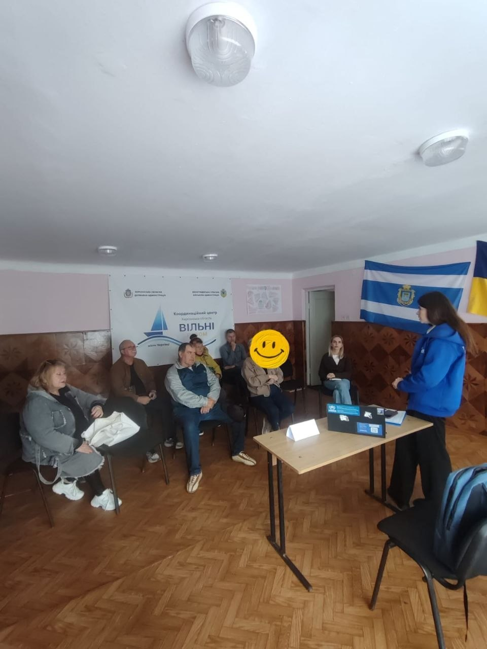 Юристка БФ «Право на захист» у синьому худі стоїть біля робочого столу з ноутбуком у Чернівцях та проводить для переселенців із Херсонщини інформаційну сесію щодо нової програми забезпечення тимчасовим житлом. Спікерка роз'яснює порядок участі у програмі групі відвідувачів, які сидять на стільцях уздовж стіни, уважно слухаючи правову інформацію. На задньому плані закріплено білий офіційний банер із синім вітрильником Координаційного центру «Вільні разом», а на стіні праворуч висять Державний Прапор України та прапор міста Херсона, що відображає процес надання безкоштовної правової та консультаційної допомоги громадянам в евакуації.