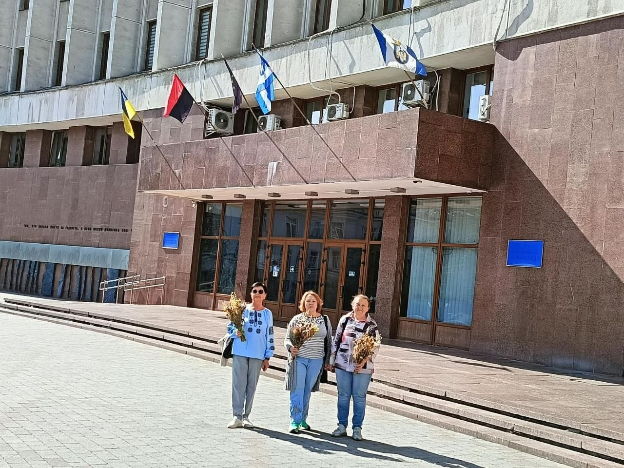 Троє жінок-переселенок із Херсонщини в повсякденному одязі стоять на площі перед головним входом до монументальної будівлі Івано-Франківської обласної державної адміністрації, тримаючи в руках флористичні композиції із сухоцвітів та колосків пшениці після творчої майстерки «Степові квіти Херсонщини». Будівля оздоблена темно-бордовим гранітом, а на флагштоках над входом майорять чотири прапори: Державний Прапор України, червоно-чорний прапор, прапор Івано-Франківська та офіційний прапор області. Жінка ліворуч одягнена у блакитну сорочку-вишиванку, дві інші учасниці — у кофти та джинси, що ілюструє проведення культурного заходу в межах проєкту «Код Херсонщини: пам’ять, культура, єдність» від центру допомоги «Вільні разом» в Івано-Франківську за підтримки Фонду громади міста Херсон «Захист» та Фонду Східна Європа за фінансової допомоги Європейського Союзу.