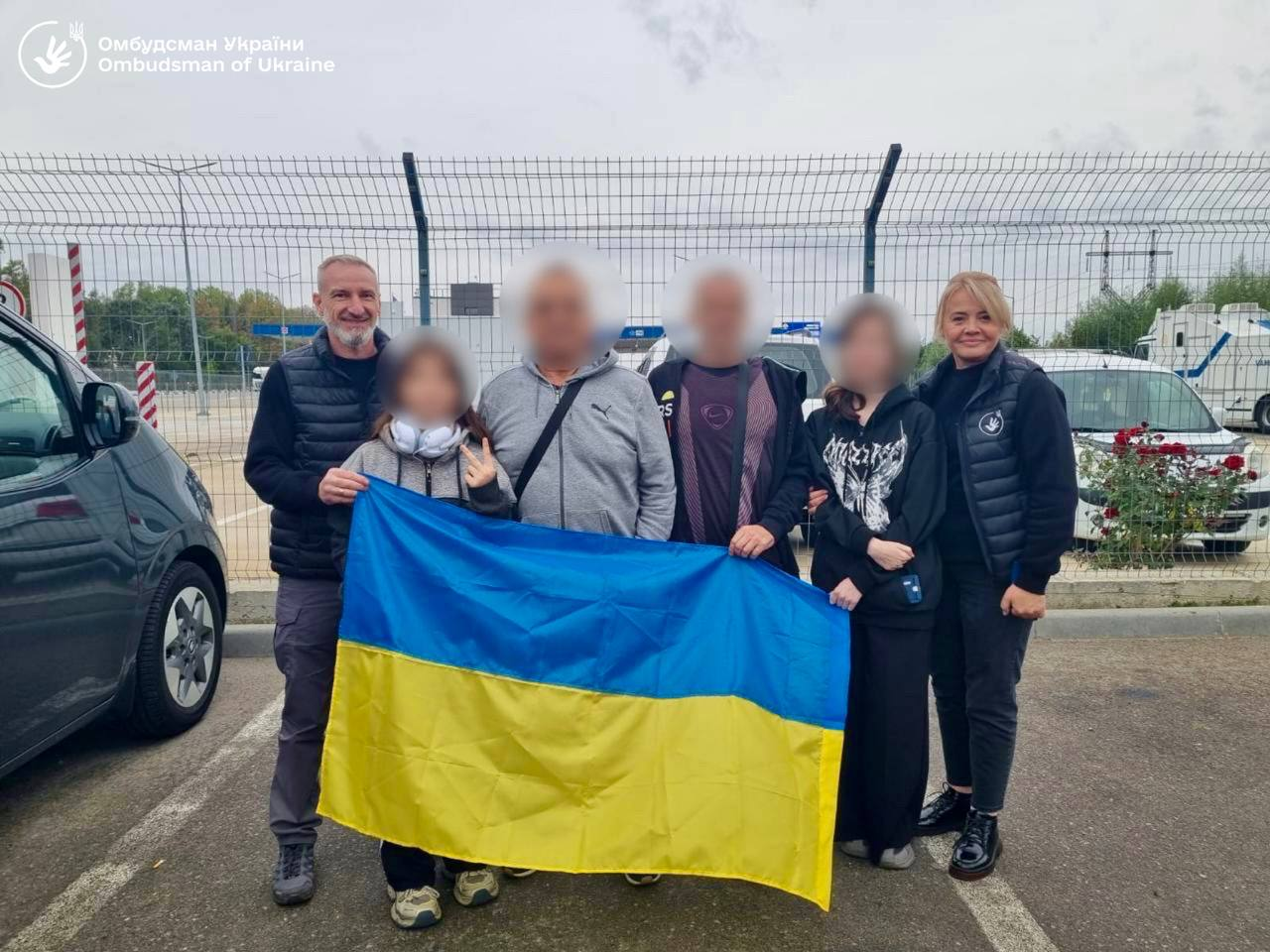 Група з шести людей, серед яких дорослі, підлітки та діти, разом тримають великий синьо-жовтий Державний Прапор України під час зустрічі на відкритому повітрі біля сітчастого металевого паркану. Ліворуч стоїть чоловік у темній жилетці, по центру розмістилися двоє чоловіків та дівчинка в навушниках, яка показує жест «V» (перемога), а праворуч позують підліток у чорному худі з малюнком метелика та усміхнена жінка. Обличчя обох підлітків та чоловіків у центрі приховані (запіксельовані) задля безпеки, а в лівому верхньому кутку кадру нанесено білий логотип і напис «Омбудсман України / Ombudsman of Ukraine», що ілюструє зусилля Офісу Омбудсмана України Дмитра Лубінця, завдяки яким вдалося повернути з тимчасово окупованих територій та через інші країни 21 українця.
