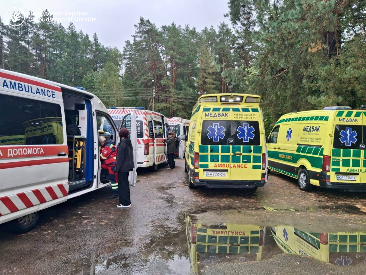 Колона евакуаційних реанімобілів та спеціалізованих автомобілів швидкої медичної допомоги («МЕДЕВАК») стоїть на дорозі під час масштабної медичної евакуації українських громадян, організованої Офісом Омбудсмана. На передньому плані ліворуч зафіксовано білий мікроавтобус із відкритими бічними дверима, біля якого працюють медики, а праворуч припарковані два жовто-зелені реанімобілі з написом «МЕДЕВАК», чиї увімкнені спецсигнали відображаються у калюжі на асфальті. У лівому верхньому кутку кадру нанесено білий логотип і напис «Омбудсман України / Ombudsman of Ukraine», що ілюструє роботу команди Дмитра Лубінця з повернення 21 українця з тимчасово окупованих територій та надання їм необхідного медичного супроводу на шляху до безпеки.