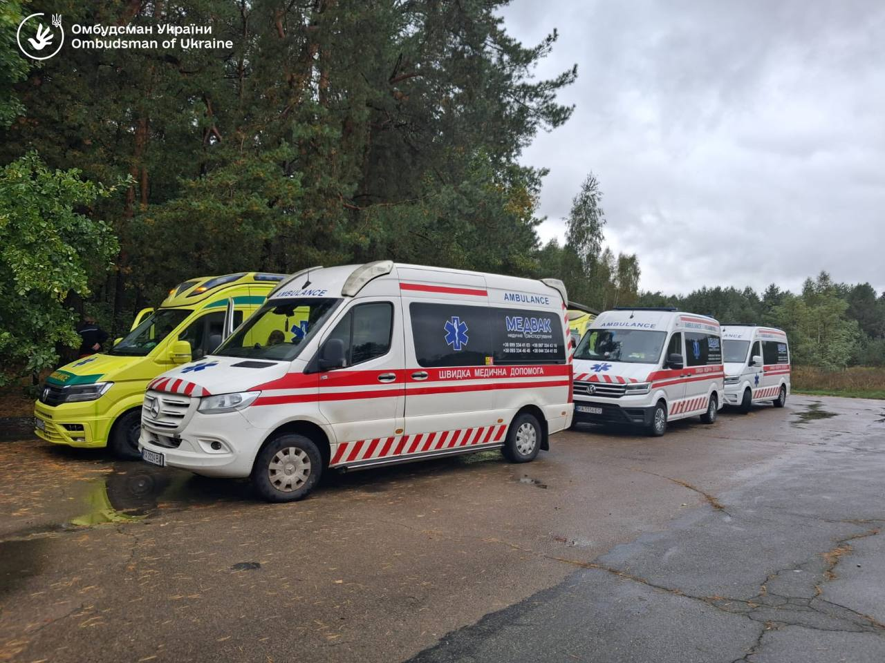 Колона автомобілів швидкої медичної допомоги та реанімобілів «МЕДЕВАК» вишикувалася на асфальтованій дорозі на тлі густих зелених дерев під час евакуаційної місії з повернення українських громадян. На передньому плані зображено білий мікроавтобус Mercedes-Benz із червоними смугами, написами «ШВИДКА МЕДИЧНА ДОПОМОГА», «AMBULANCE» та емблемою «Зірка життя». Позаду нього в один ряд стоять ще кілька спецавтомобілів, серед яких виділяється яскраво-жовтий реанімобіль ліворуч, а небо над ними затягнуте хмарами. У лівому верхньому кутку кадру розміщено білий логотип та напис «Омбудсман України / Ombudsman of Ukraine», що відображає роботу команди Дмитра Лубінця з евакуації 21 українця з тимчасово окупованих територій, де через складний стан здоров'я повернутих родин із Херсонщини було забезпечено повний медичний супровід та координацію з гуманітарними організаціями.
