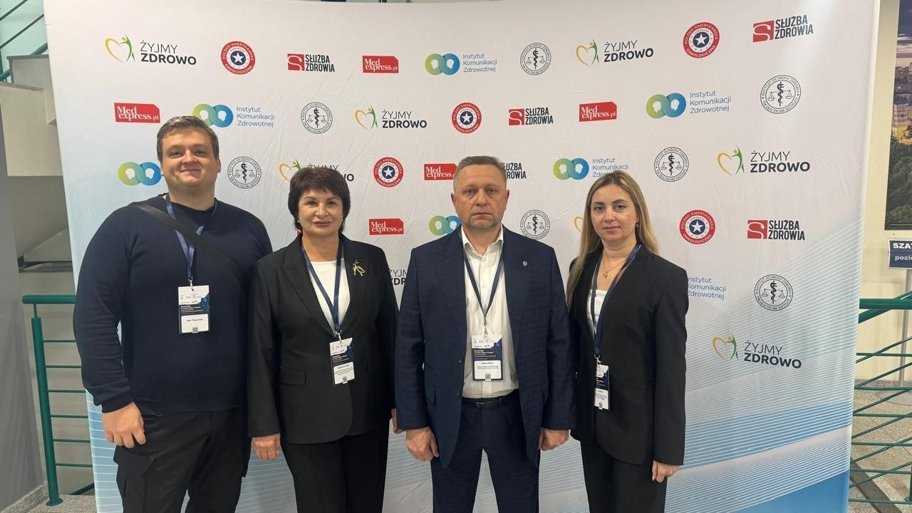 Чотири представники делегації Херсонської області у класичних темних костюмах та повсякденному одязі з офіційними бейджами на шиї стоять у ряд та позують на тлі великого світлого банера з чисельними медичними логотипами під час масштабного міжнародного заходу у Варшаві. Зліва направо на знімку зафіксовані: головний спеціаліст управління міжнародного співробітництва та залучення інвестицій Херсонської ОДА Іван Ткаченко, заступниця начальника Херсонської МВА Наталія Чорненька із жовто-блакитною стрічкою на лацкані піджака, перший заступник начальника Херсонської ОВА Дмитро Бутрій у темно-синьому піджаку та заступниця директора установи «Агенція регіонального розвитку «Офіс євроінтеграції» Херсонської області» Інна Бутрій. Кадр ілюструє участь посадовців у безпековій конференції «Місцеве самоврядування в умовах війни та кризи: уроки українського досвіду», де херсонська делегація презентувала польським колегам та урядовцям протоколи кризового адміністрування, безпекові рішення для прифронтових громад, розвиток мережі підземних лікарень і шкіл, а також роботу координаційних центрів підтримки ВПО «Вільні разом».