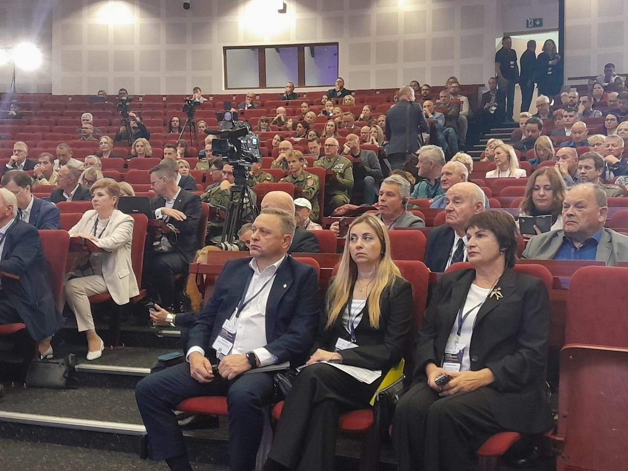 Представники делегації Херсонської області зосереджено сидять у першому ряду великої амфітеатральної зали конференції у Варшаві серед чисельних іноземних учасників, експертів та військових під час обговорення кризового адміністрування прифронтових територій. На передньому плані на червоних кріслах поруч сидить керівна ланка херсонської делегації: перший заступник начальника Херсонської ОВА Дмитро Бутрій у темно-синьому костюмі й білій сорочці, заступниця директора установи «Агенція регіонального розвитку «Офіс євроінтеграції» Інна Бутрій у чорному брючному костюмі та заступниця начальника Херсонської МВА Наталія Чорненька із золотистою стрічкою-тризубом на лацкані піджака. Позаду них усі ряди великого залу заповнені сотнями слухачів, дипломатів, представників екстрених служб та польських посадовців, а по центру зали між кріслами встановлено професійні телевізійні камери на штативах, що фіксують презентацію українського безпекового досвіду, створення підземних лікарень та антидронових загороджень на деокупованих територіях.